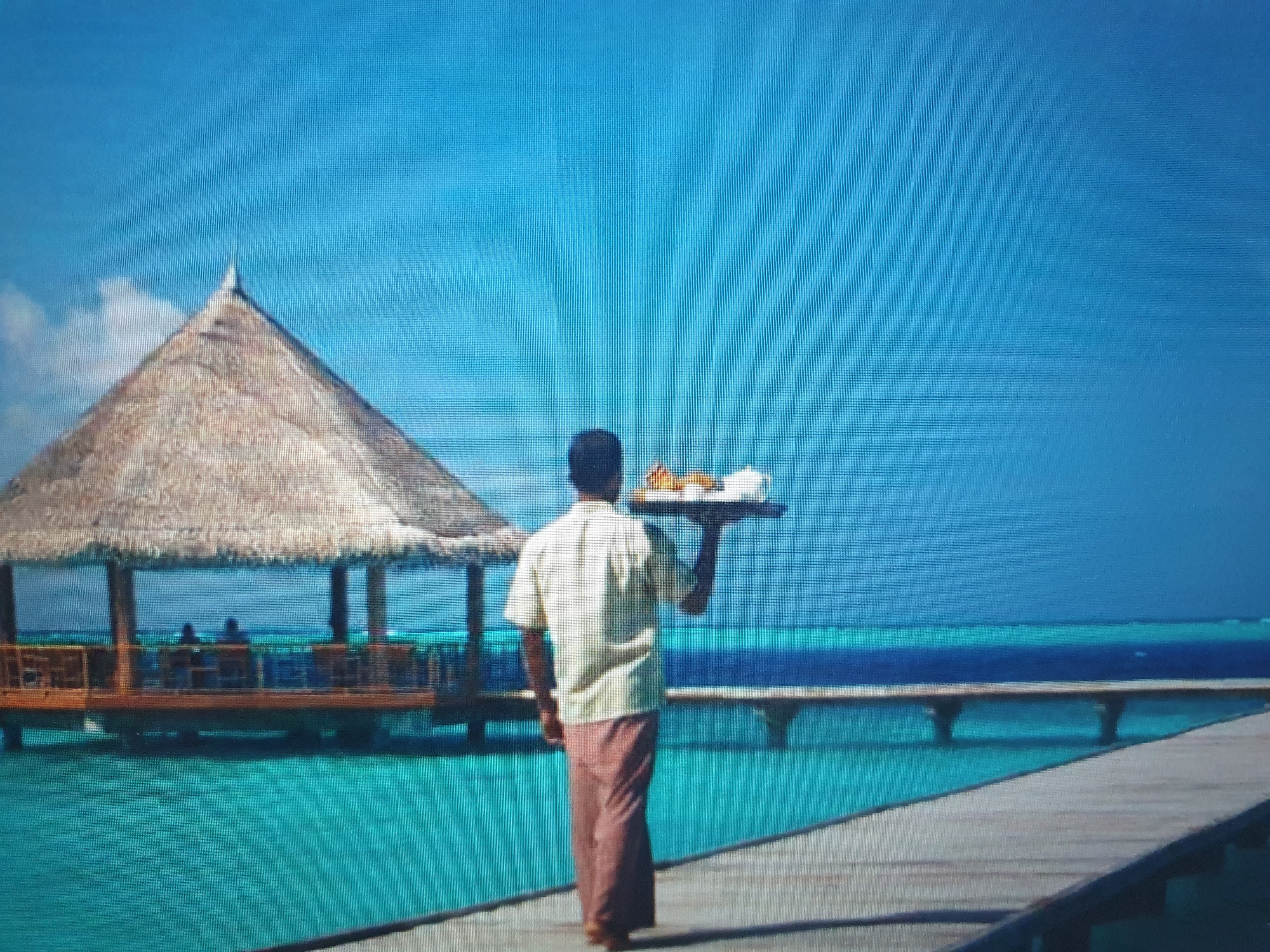 The image depicts a person in a light-colored shirt and brown trousers carrying a tray of food, walking on a wooden pier over turquoise water. To the left, there is a thatched-roof structure resembling a gazebo or shelter on stilts, with people visible inside. In the background, a bridge or walkway extends over the water, and the horizon shows the transition from the turquoise water to a deep blue sky with some light clouds. The scene is set in a tropical or subtropical environment, likely a resort area with overwater structures. There is no indication that this scene is located in the city of Tashkent, Uzbekistan, which is an inland city with a different climate and landscape.