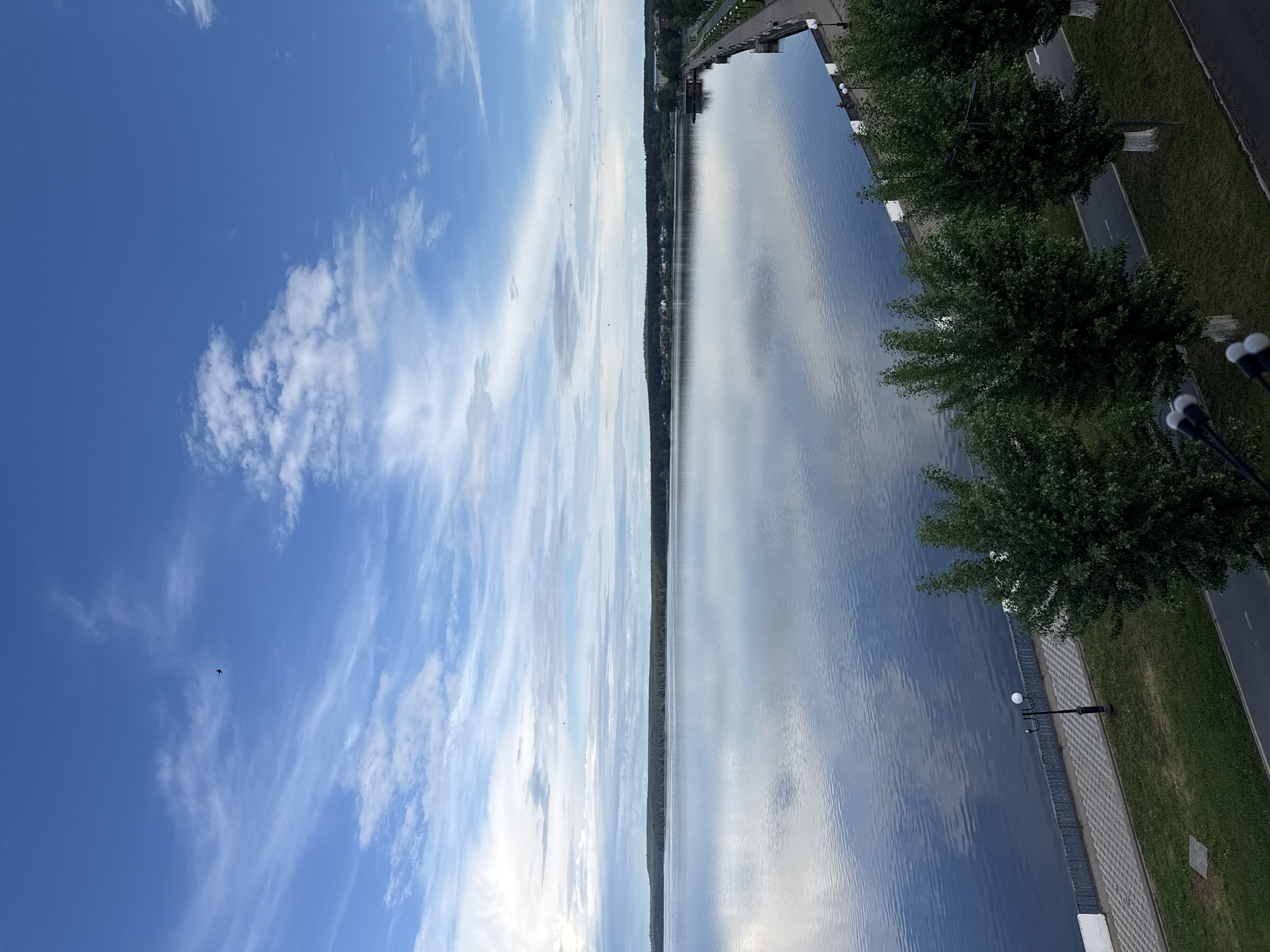 The image shows a body of water, possibly the Votkinsk Reservoir, a prominent feature of Votkinsk, Russia. The sky, filled with clouds, is reflected on the water surface. Along the right side of the frame, there's a walkway with trees and street lights, suggesting a park or promenade area. The opposite shore is lined with trees.