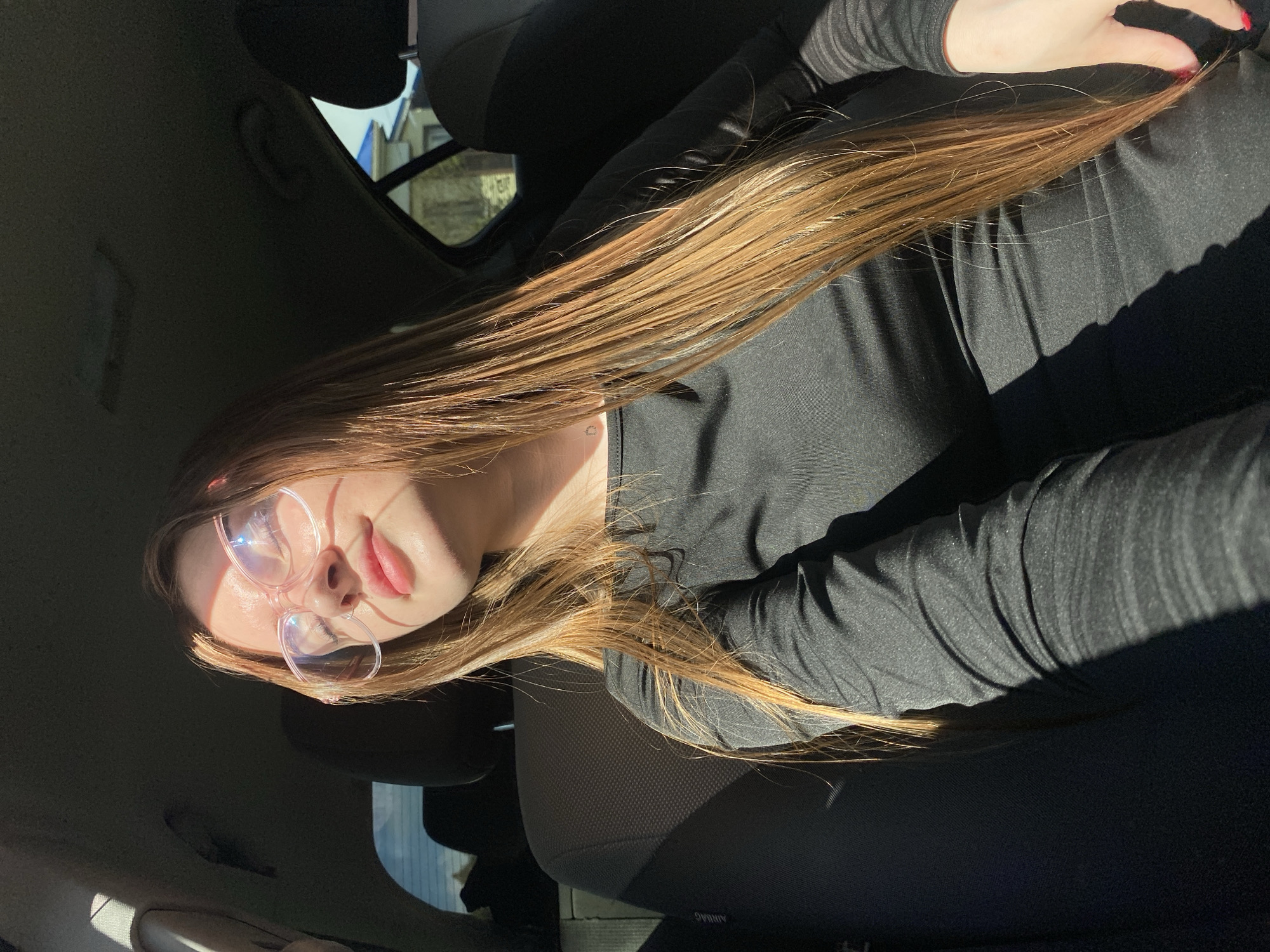 A young woman with long, light brown hair and pink-rimmed glasses is sitting in the passenger seat of a car. Sunlight streams through the window, illuminating her face and hair. She is wearing a black long-sleeved shirt. The interior of the car is visible, including the headrest of the driver's seat and a portion of the backseat. Outside the car window, a residential building with a blue roof and trees can be seen, suggesting a suburban or rural setting, potentially in the village of Dyakovo, Russia. The image is captured from a slightly elevated angle, looking down at the woman.