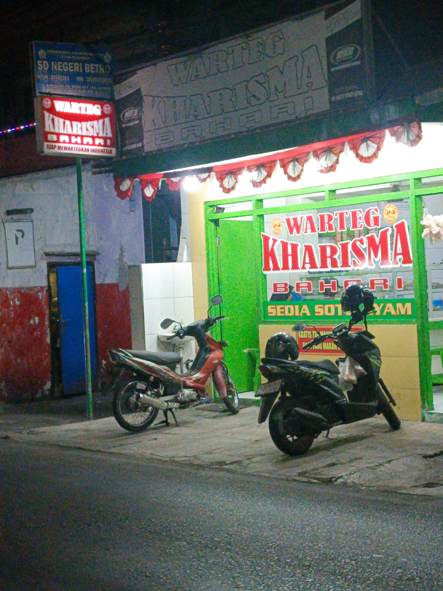 In Sedati Agung, Indonesia, a brightly lit "WARTEG KHARISMA BAHARI" restaurant stands on a street at night. Two motorcycles are parked outside the restaurant. One is a red and white scooter, and the other is a black scooter with two helmets on its handlebars. The restaurant sign, illuminated in red and white, features the text "WARTEG KHARISMA BAHARI" and "SEDIA SOT YAM". Red and white bunting hangs above the entrance. A blue door is visible on an adjacent building, which also has a sign for "SD NEGERI BETRO". The street is paved with asphalt, and the surrounding buildings appear to be typical of a commercial area in Indonesia.