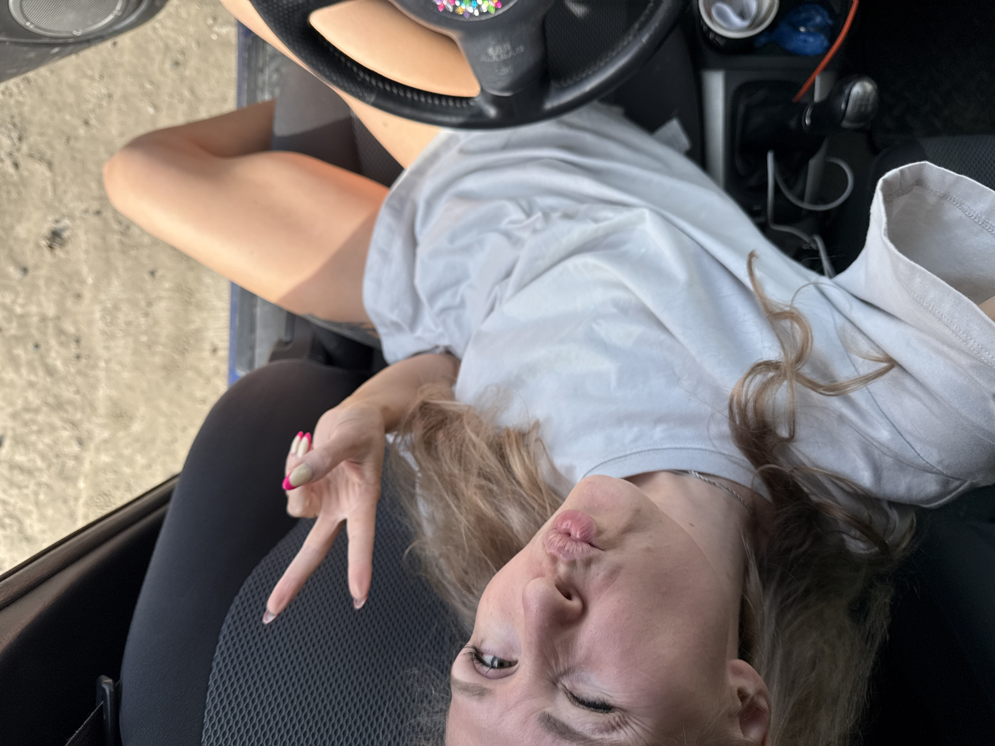 A woman is seated in the driver's seat of a car. She is wearing a light-colored t-shirt. She is holding up two fingers in a peace sign with her right hand. She is making a kissing face and winking one eye. The car's steering wheel is visible in the foreground. The location is the city of Chaikovsky, Russia. The car's interior includes a gear shift and cupholders. Through the open car door, a sandy surface is visible.