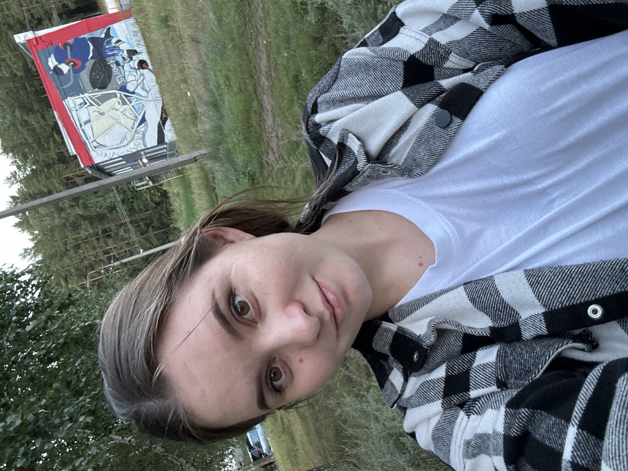 In Noviy, Russia, a young woman with brown hair and brown eyes is captured in a selfie taken outdoors. She is wearing a white t-shirt and an open black and white plaid shirt. The background reveals a grassy area with trees and a building adorned with graffiti depicting a hockey scene. The sky is overcast, suggesting it might be late afternoon or early evening.