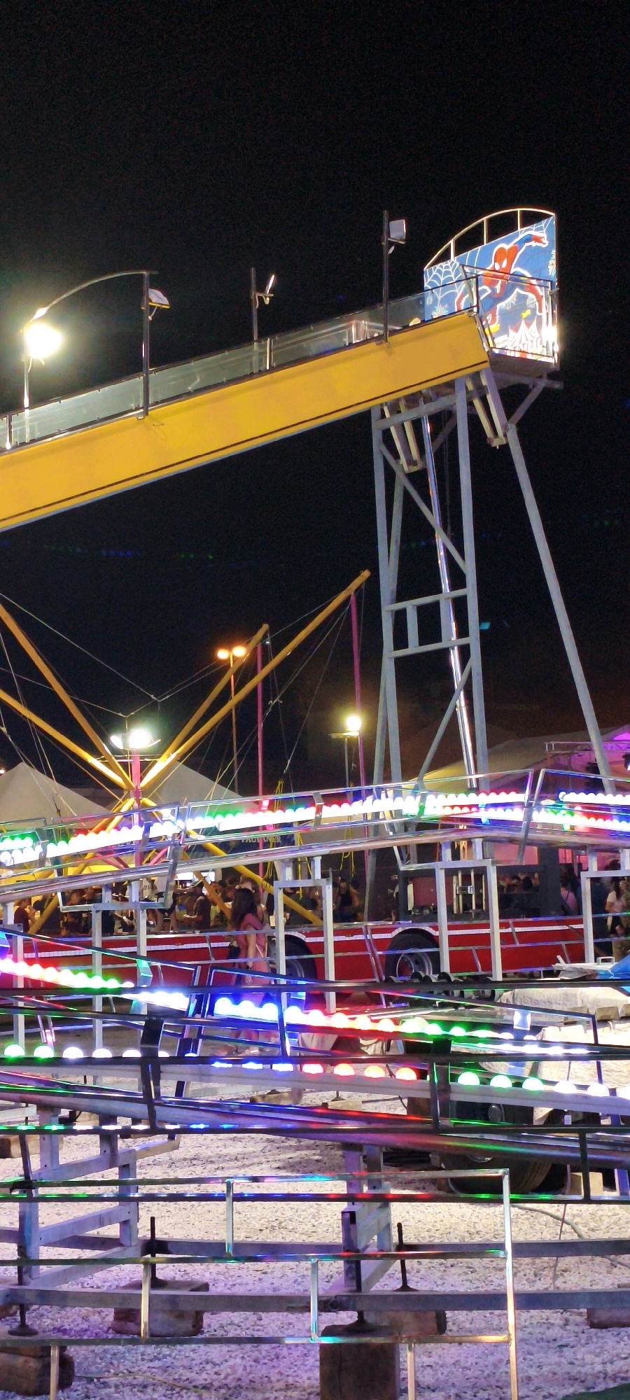 This is a nighttime photograph taken at a fair in Campiglia Marittima, Italy, possibly during the "Palio di San Giovanni Decollato" festival. The scene features amusement park rides, illuminated with vibrant multi-colored lights, primarily red, green, blue, and white. In the foreground, there is a complex structure of metal railings and supports, likely part of a ride's track or queue. Behind this, a red, bus-like amusement ride is visible, crowded with people. The ride is outlined with strings of colorful LED lights. Further in the background, the yellow ramp of another attraction extends upwards against a dark sky. There are also tent structures and light poles visible, contributing to the festive atmosphere. The ground appears to be covered with gravel. The overall impression is of a lively and colorful evening at a fairground.