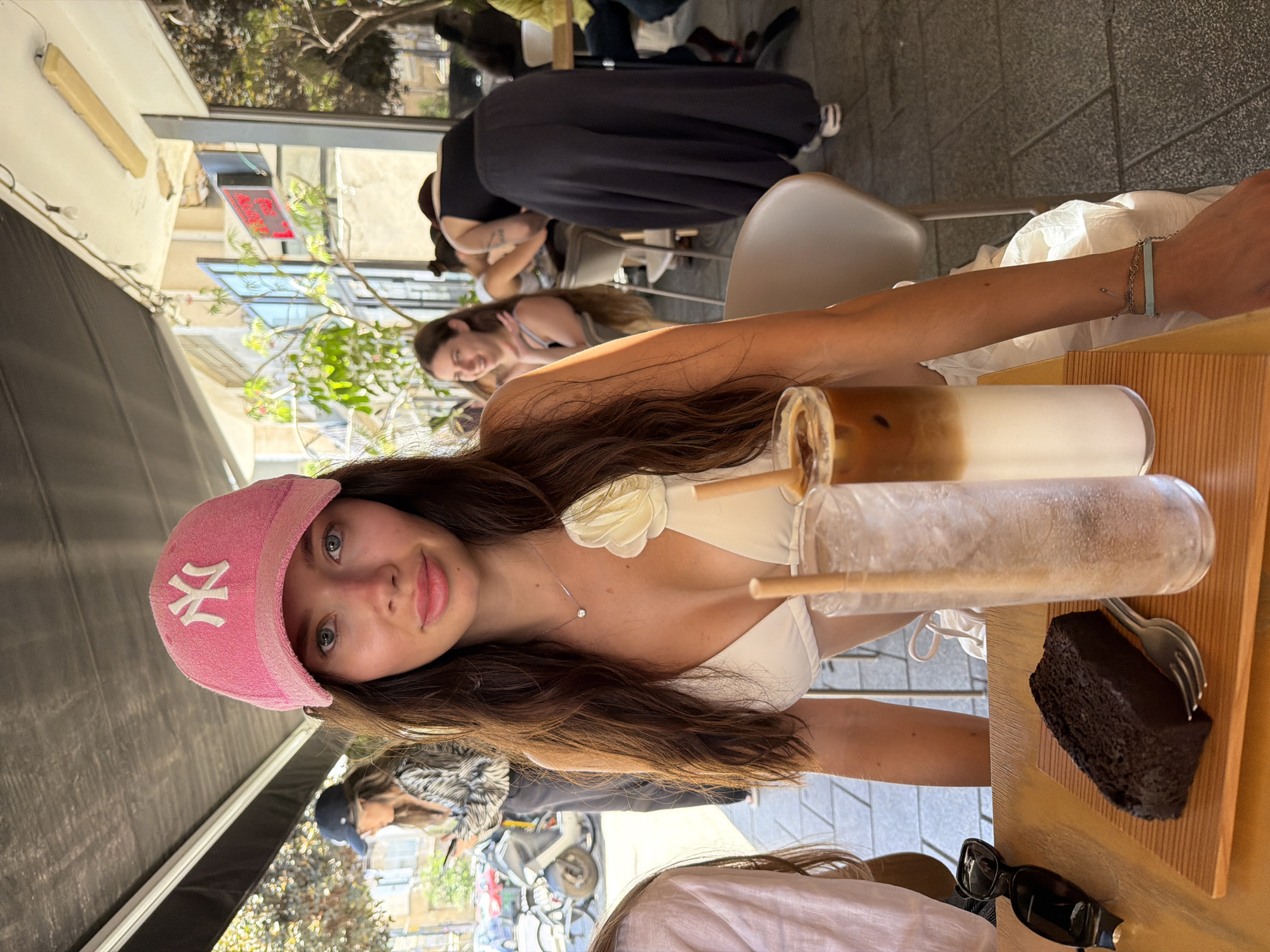 A young woman with long, dark brown hair, wearing a pink terry cloth New York Yankees baseball cap, is seated at an outdoor cafe table, looking directly at the camera. She wears a white bikini top with a white floral embellishment on the right strap and a silver necklace with a single round pendant. Her left forearm rests on the table, displaying a light blue bar-shaped bracelet. On the wooden table, a ridged wooden board holds a rectangular dark brown cake or bread slice and a silver fork. Two tall glasses are present: one with iced water and a straw, and another with a layered iced coffee. Black sunglasses are positioned to the left of the wooden board.
In the mid-ground to the left, another woman wearing a dark cap is partially visible, focusing on a mobile phone. Behind her, a street scene features parked scooters and multi-story building facades. To the right of the main subject, a woman with long hair is seated at a table, smiling toward the right. Further back, a woman in a black sleeveless top and dark, loose-fitting pants is bent over, her back to the camera. The cafe operates under a dark awning with exposed light bulbs. A red digital display is visible on a building across the street. The urban setting, characterized by outdoor cafes and local architecture, is consistent with the city of Tel Aviv, Israel.