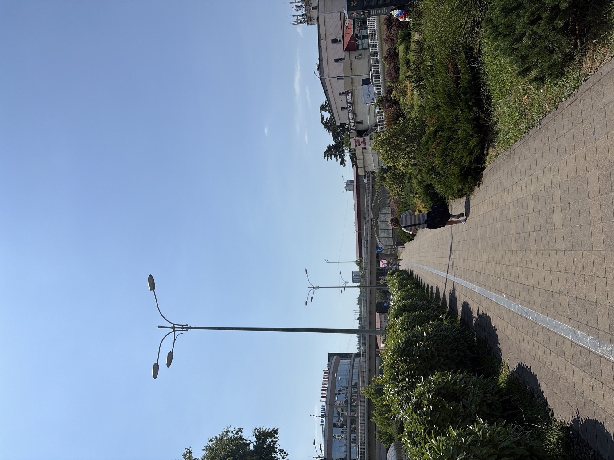The image depicts a sunny day on a pedestrian walkway in Sochi, Russia. The walkway is paved with tan-colored rectangular tiles and runs alongside a neatly trimmed green hedge. A person, seen from behind, is walking away from the camera on the walkway. This individual is wearing a striped t-shirt, dark shorts, and carrying a dark bag over their right shoulder. In the background, a multi-story building with large windows is visible, with Russian text above it reading "ТОРГОВЫЙ ЦЕНТР ГАГАРИНСКИЙ" (Gagarinskiy Shopping Center). Beyond the shopping center, an elevated road or bridge structure crosses the scene, with streetlights lining its edge. Further in the distance, other buildings and trees can be seen under a clear blue sky. The scene suggests an urban environment with commercial and public spaces.