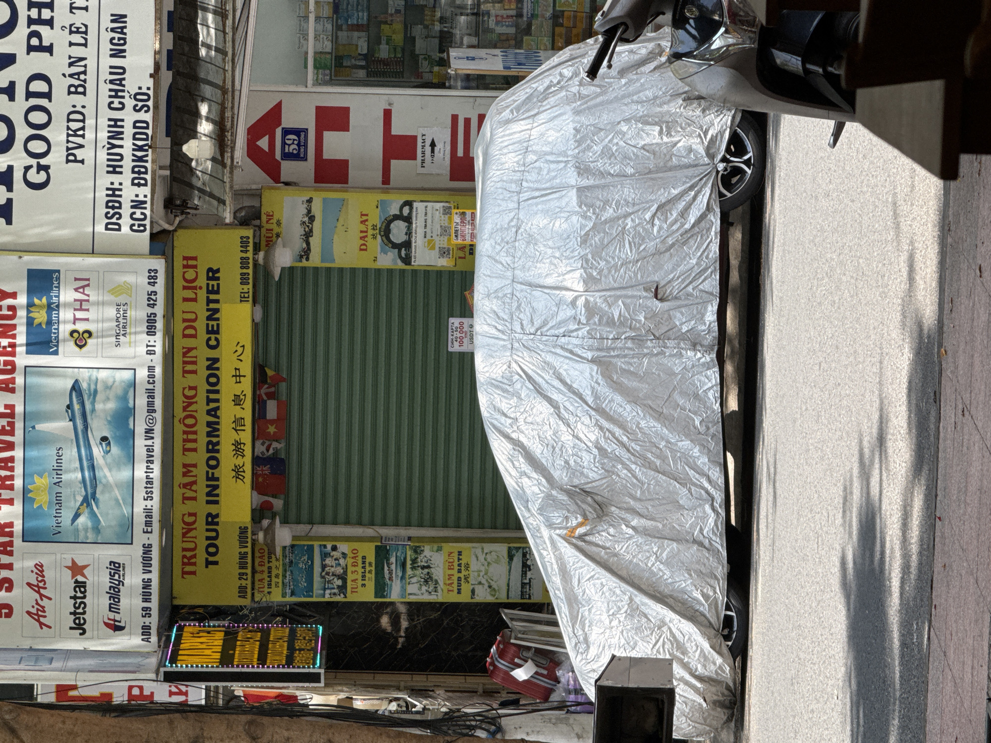 The image captures a street scene in Nha Trang, Vietnam, likely in the vicinity of tourist-oriented businesses. In the foreground, a car is covered by a large, wrinkled silver tarp, with a portion of a scooter visible on the right. The tarp obscures the car's details, but a wheel with a distinctive spoke pattern is visible. The background is dominated by shopfronts with various signage.
A prominent yellow sign reads "TRUNG TÂM THÔNG TIN DU LỊCH TOUR INFORMATION CENTER" in Vietnamese and English, along with Chinese characters. Below this, a sign advertising "TUA 3 DAO 3 ISLAND TOUR" and "TÂM BÙN MUD BATH" is displayed vertically with images of activities and locations. Other signs feature airline logos such as Air Asia, Jetstar, Malaysia Airlines, Vietnam Airlines, Thai Airways, and Singapore Airlines, suggesting travel agencies. Some signs also display addresses and phone numbers. A red sign with a white arrow and the number "59" is also visible, likely indicating a building number. A pharmacy sign with the word "PHARMACY" and an arrow is present on the right side of the frame. The overall impression is of a commercial area catering to tourists in Nha Trang.