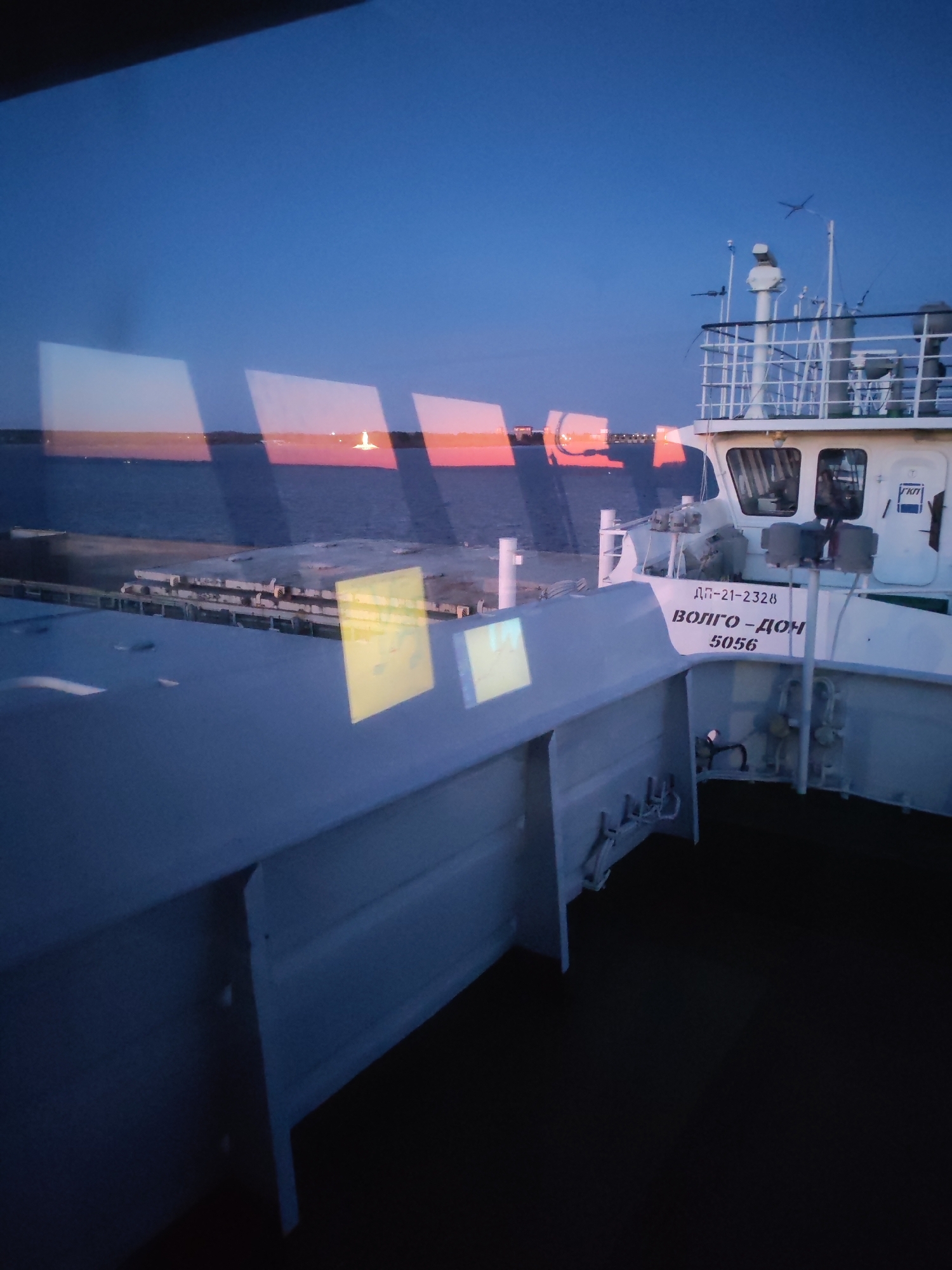 The image depicts a twilight or dawn scene viewed from inside a vessel, looking out through a window with prominent reflections. Rectangular patches of light in yellow, pink, and orange, originating from the interior, are reflected on the glass, partially obscuring the view of the outside environment.
To the right, a white, modern vessel is visible, likely a cargo or passenger ship. Its side features distinct text: "ДП-21-2328", "ВОЛГО-ДОН" (Volgo-Don), and "5056". The vessel has a prominent bridge/cabin area with windows, railings, and a mast equipped with radar or communication apparatus.
Beyond the immediate foreground, a wide body of calm, dark water extends towards the horizon. In the mid-distance, a dark strip of land or industrial structures is discernible. Against the horizon, where the sky transitions from a deep blue to a soft pink-orange glow, a distinct, brightly illuminated white structure, possibly a lighthouse or a monument, stands out. The sky above is a clear, dark blue, indicating good weather. Part of the white structure of the vessel from which the photo is taken is visible in the lower foreground.
The scene captures a peaceful moment on a waterway, likely near Rybinsk, Russia, given the "Volgo-Don" inscription, which refers to the famous canal system. No individuals are visible in the image.