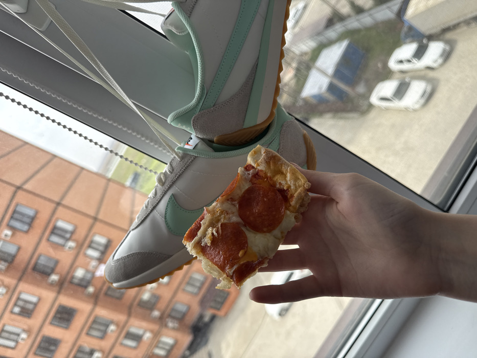 A human hand in the lower-left foreground holds a single slice of pepperoni pizza, featuring a thick crust, melted cheese, and several circular pepperoni pieces. Directly behind and above the pizza, a pair of new white, light gray, and mint green Nike athletic shoes hang by their white laces from an unseen fixture above a window. Through the window, an urban residential area is visible under an overcast sky. A prominent multi-story building constructed of orange-brown brick, with numerous windows and exterior air conditioning units, occupies the mid-ground. Below, a paved surface features two parked white sedans, and a grassy area contains two blue tarp-covered structures. The scene is observed from an elevated indoor position, looking out onto a street or courtyard in an Unknown city, Unknown region.
