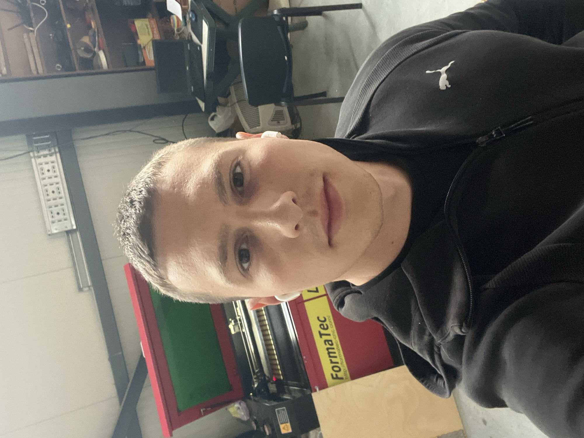 A selfie taken by a light-skinned male with short, dark hair and wearing a black Puma-branded zip-up jacket is positioned in the foreground. He has an earbud in his ear. Behind him is a Formatec CNC machine, with a red frame and a green-tinted window. Part of a tan wood panel is visible near the CNC machine.
In the background, to the left, is a white wall with a gray metal support structure and a white electrical panel. Further back, a shelf with various items is discernible. A black office chair is also visible.
The image does not contain enough information to determine a specific location within Burgas, Bulgaria.