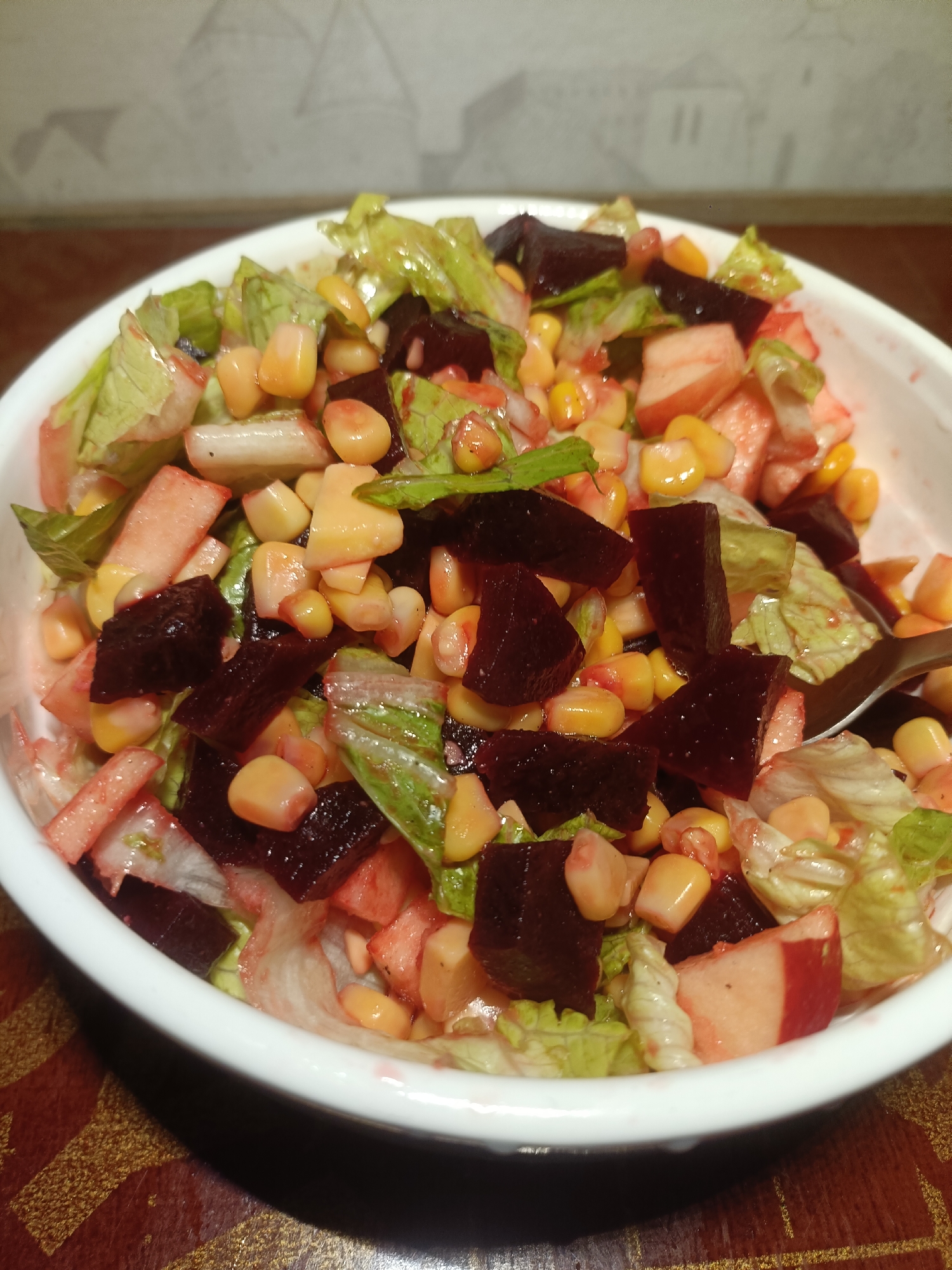 The image shows a white bowl filled with a salad. The salad contains lettuce, corn, diced beets, and what appears to be chopped fruit, possibly apples. A spoon is visible within the salad. The bowl is placed on a surface with a patterned design, possibly a table. The background shows a wall with a patterned wallpaper. There is no indication that the image was taken in Dubai, United Arab Emirates.