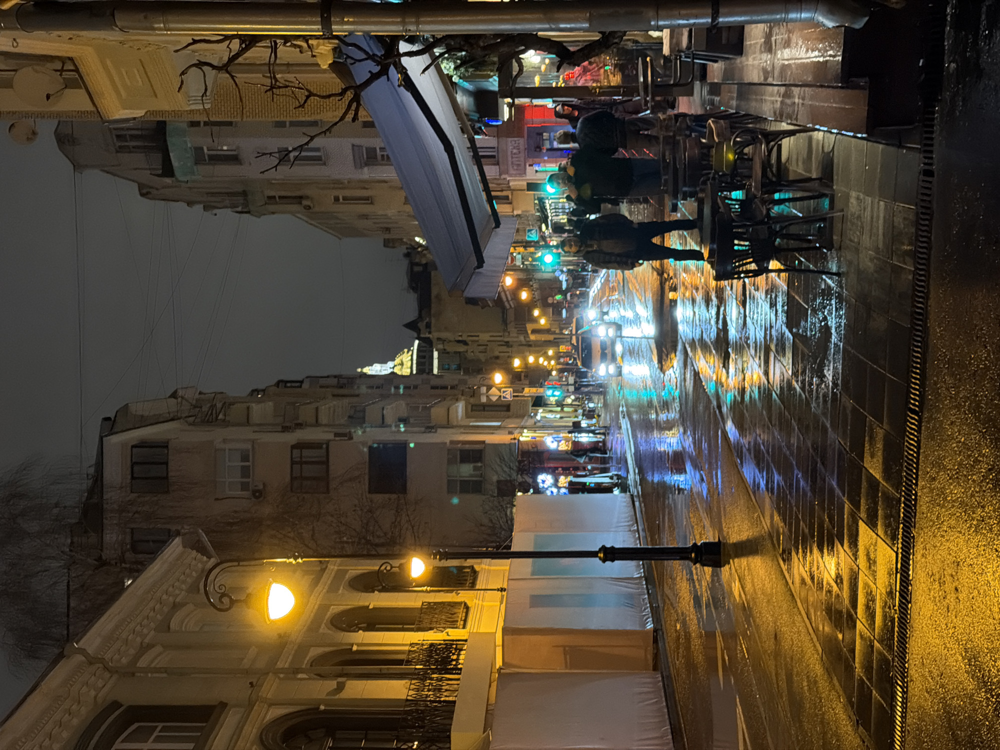 The image captures a nighttime street scene in Moscow, Russia, under wet conditions, likely after rainfall. The street is lined with buildings of varying heights and architectural styles. On the right side, a sidewalk with dark, reflective tiles leads past an outdoor cafe. Several people are visible near the cafe, some sitting and some standing. Streetlights and building lights create reflections on the wet pavement, creating a vibrant display of colors. A street lamp is visible in the foreground on the left side of the road. Overhead power lines stretch across the sky. The Russian word "APTEKA" is prominently displayed on a storefront. The overall atmosphere is urban and lively, despite the rain.