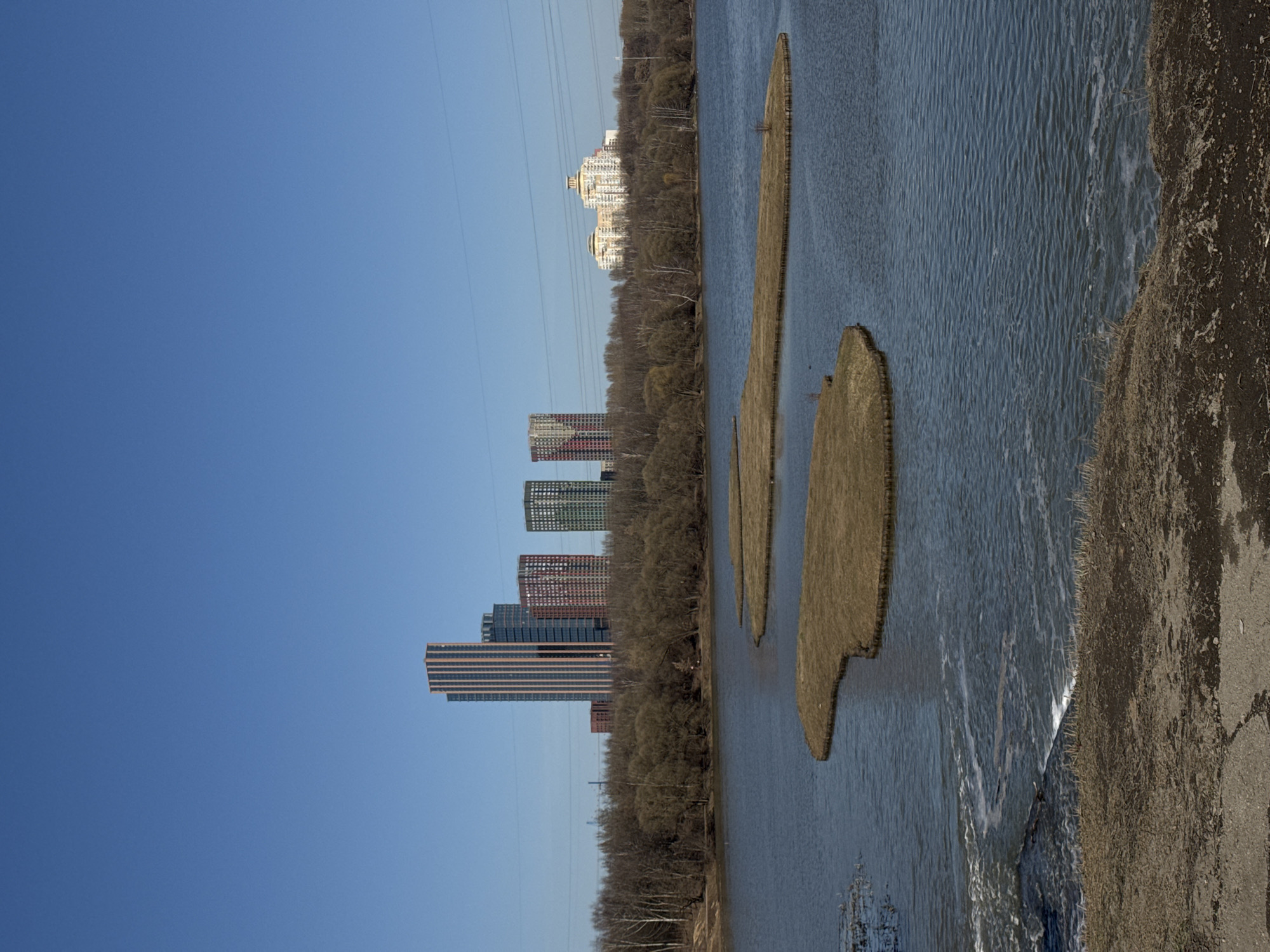 The image depicts a river landscape in Moscow, Russia. A series of modern high-rise buildings line the left bank, while the right bank is characterized by vegetation. Three elongated, grassy islands are visible within the river. A concrete embankment is present on the right side of the frame. The sky is a clear blue, and power lines are visible stretching across the scene.