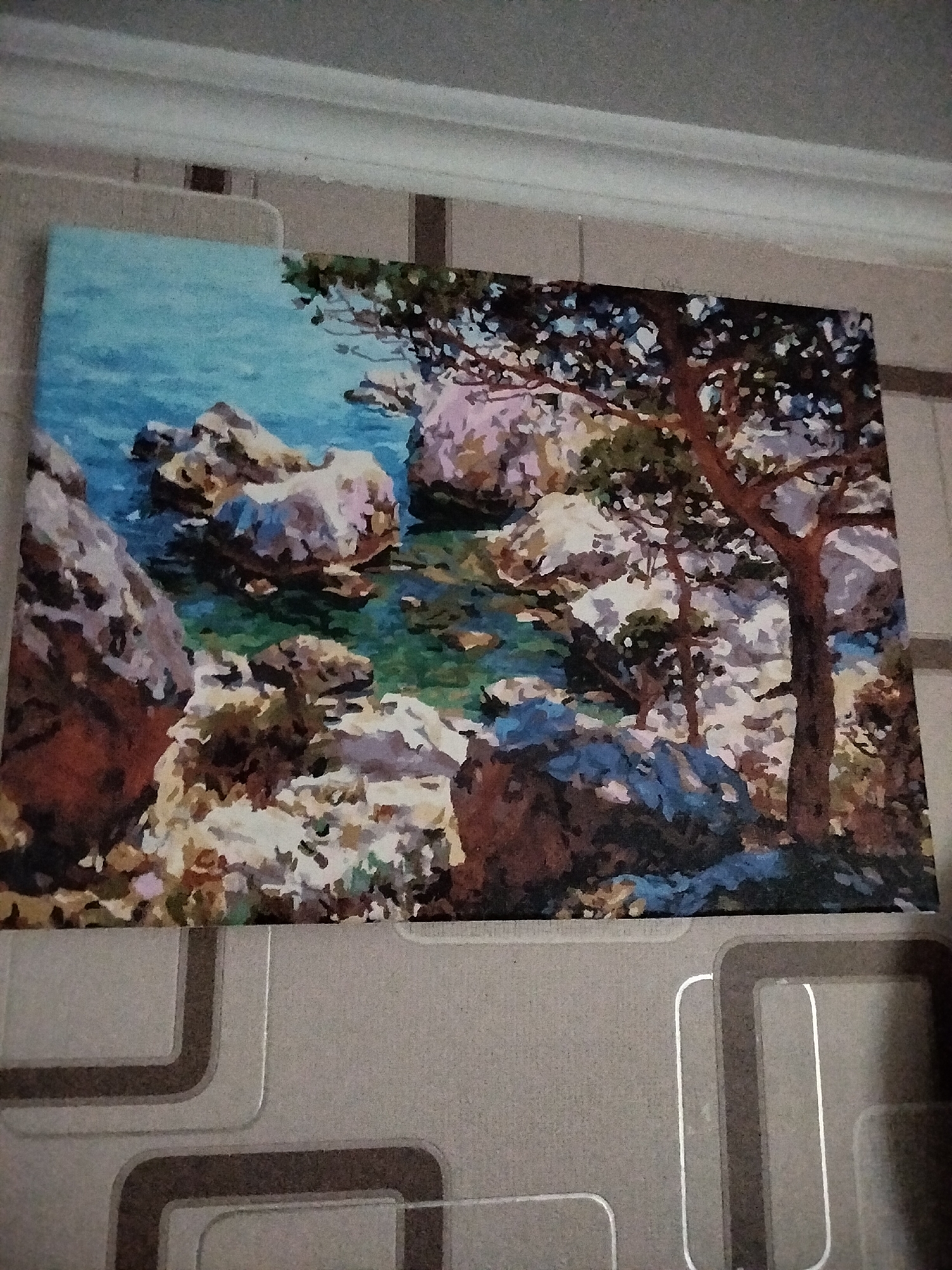 This is a photograph of a painting hanging on a wall. The painting depicts a rocky coastline with a tree in the foreground. The water is a vibrant turquoise and green, and the rocks are a mix of brown, grey, and white. The tree has a thick, brown trunk and dark green foliage. The background appears to be a blue sky. The painting is in a somewhat abstract style, with bold brushstrokes. The wall itself is textured and appears to be painted in a neutral color, with a decorative pattern of squares and lines. The painting is illuminated by artificial light, casting a slight glare on its surface. There are no people visible in the image. It is not possible to determine the location of Nizhnedonskoy, Russia from the visual information provided.