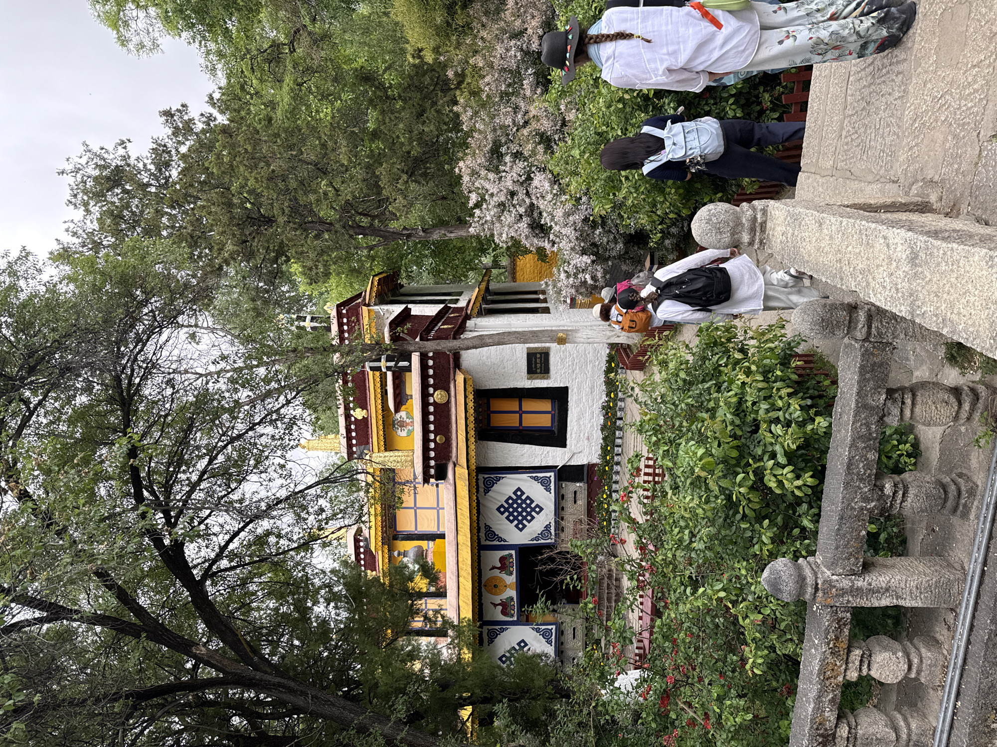 The image depicts a white and yellow building with intricate patterns and ornate decorations, located in the Chengguan District of China. The building has multiple levels and windows. Some people are seen walking on a stone path or bridge near the building. The surrounding environment consists of lush greenery, including trees and bushes. The sky appears overcast.