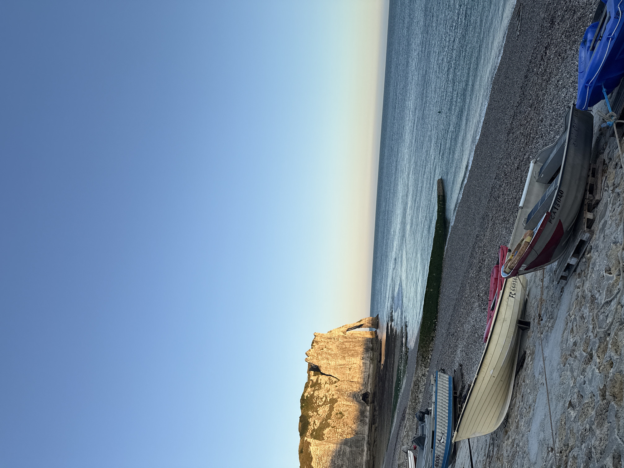 The image depicts a serene coastal scene in Étretat, France, during what appears to be either sunrise or sunset. The iconic white cliffs of Étretat dominate the left side of the frame, catching the warm, golden light of the low sun. An archway is visible carved into the cliff face. The sea stretches out to the right, its surface calm and reflecting the sky's gradient from a clear blue overhead to a softer hue near the horizon.
In the foreground, a pebbled beach transitions to a concrete embankment. Several boats are resting on the shore. One prominent light-colored rowboat, with the registration "FC 660560" visible, is positioned on a concrete ramp. Behind it, a darker-colored boat with red and white detailing is partially visible, also on wooden supports. Further up the beach, a blue boat with a white trim is tied with rope. Other boats, some with outboard motors, are visible further in the background, along the shoreline. The overall impression is one of quiet repose, with the natural beauty of the cliffs and sea contrasting with the presence of human activity through the moored boats. No people are visible in the scene.