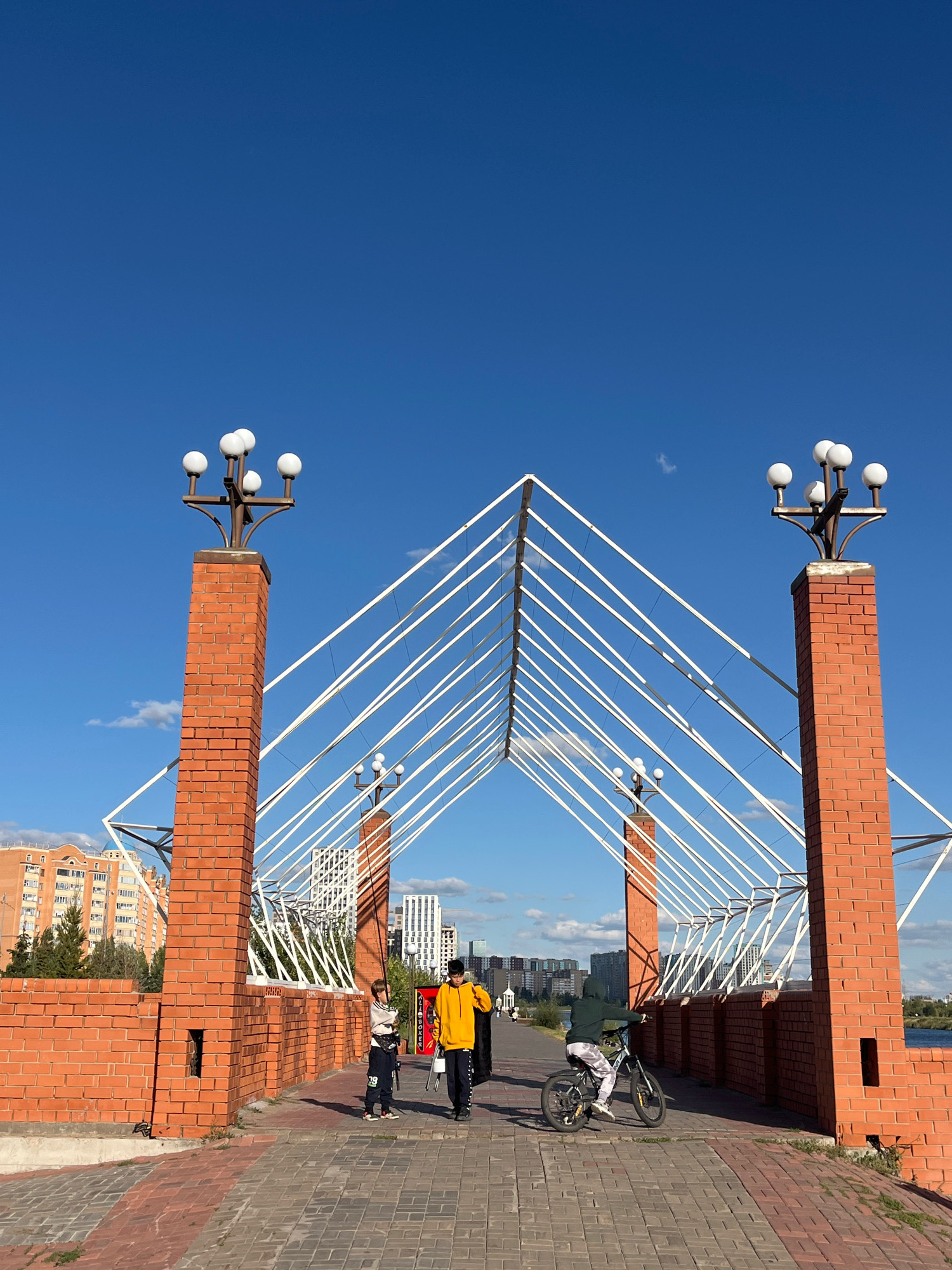 The image captures a view of the pedestrian bridge in Astana, Kazakhstan, on a clear day with a bright blue sky. The bridge features two prominent brick pillars with globe-shaped lamps on top. A distinctive white, tent-like structure made of thin cables forms the overhead canopy of the bridge. The walkway is paved with brick pavers.
In the foreground, three individuals are present. Two young boys, one in a yellow hoodie and the other in a dark green hoodie, are walking on the bridge. The boy in the green hoodie is riding a bicycle. A third, smaller child in a white and dark patterned shirt is walking ahead of them. In the background, modern city buildings of various heights and architectural styles are visible. The environment suggests an urban park or recreational area adjacent to a body of water, likely the Ishim River, given the location.