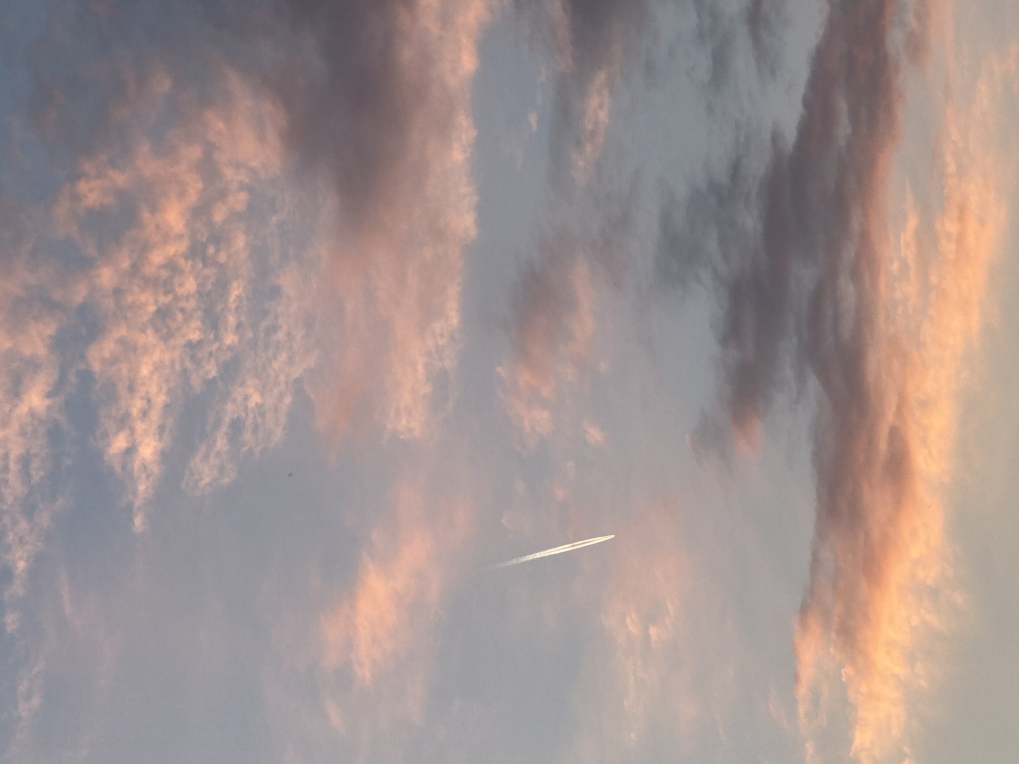 A vertical, full-frame shot captures a dusky sky with scattered clouds tinged with pink and orange hues from the setting sun. A white contrail from an aircraft streaks diagonally across the frame from the upper left to the lower right, subtly breaking the otherwise tranquil scene. The clouds are varied in density and texture, with some appearing wispy and translucent, while others are more substantial and shadowed, suggesting different altitudes and formations. The overall lighting is soft and diffused, indicative of twilight conditions. The location is unknown.