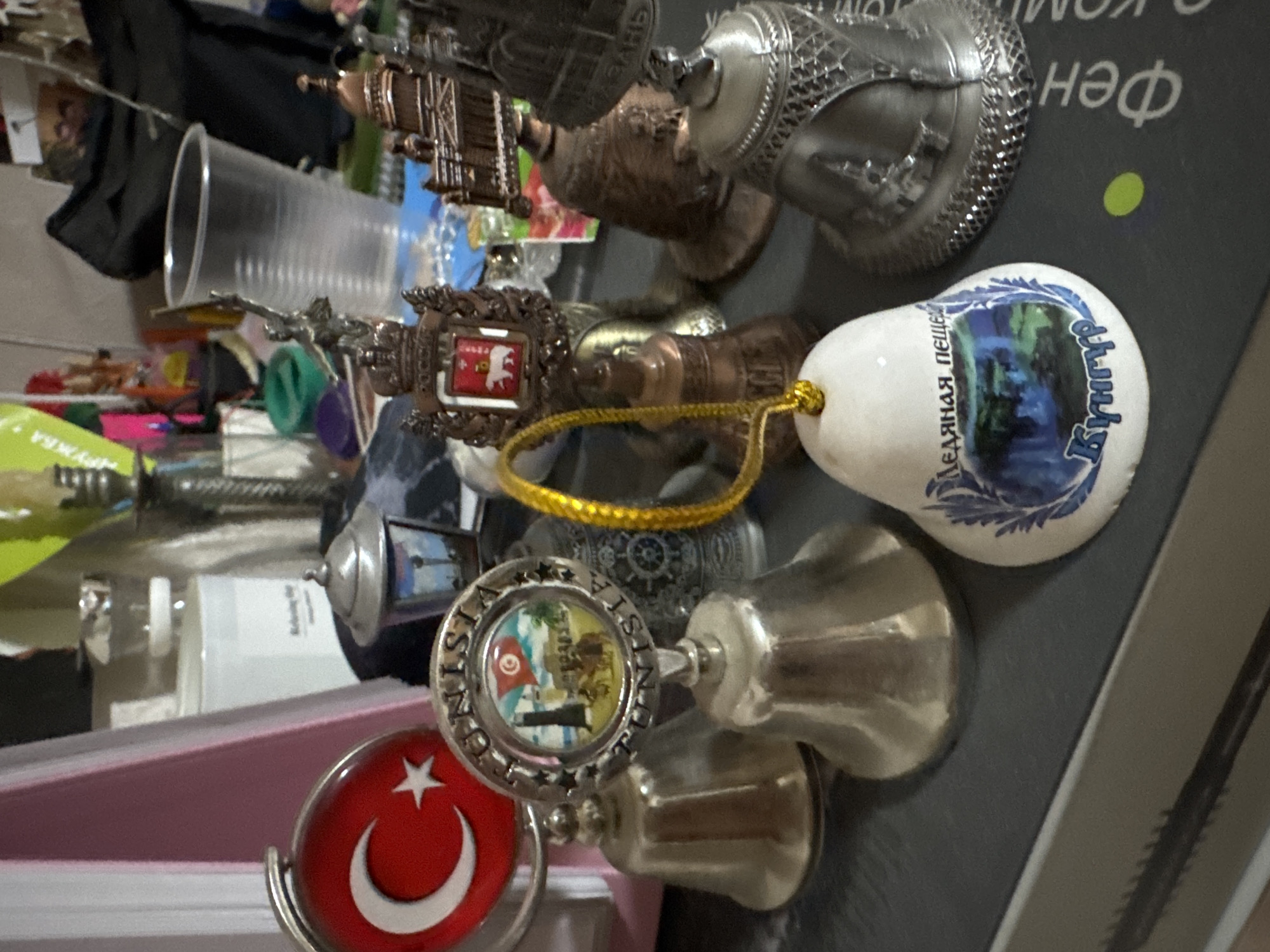 The image shows a collection of souvenir bells, likely on a shelf or table. In the foreground, a white bell with blue decorative elements and Russian text is visible. To the left, several metallic souvenir bells are arranged, including one with a Turkish flag motif and another with a scene that appears to be from Tunisia. In the background, a clear plastic cup and various other decorative items and containers are present. The scene is dimly lit, suggesting an indoor setting. The presence of Russian text on one of the bells indicates a connection to Russia, and given the provided context, this collection could be located in Izhevsk, Russia.