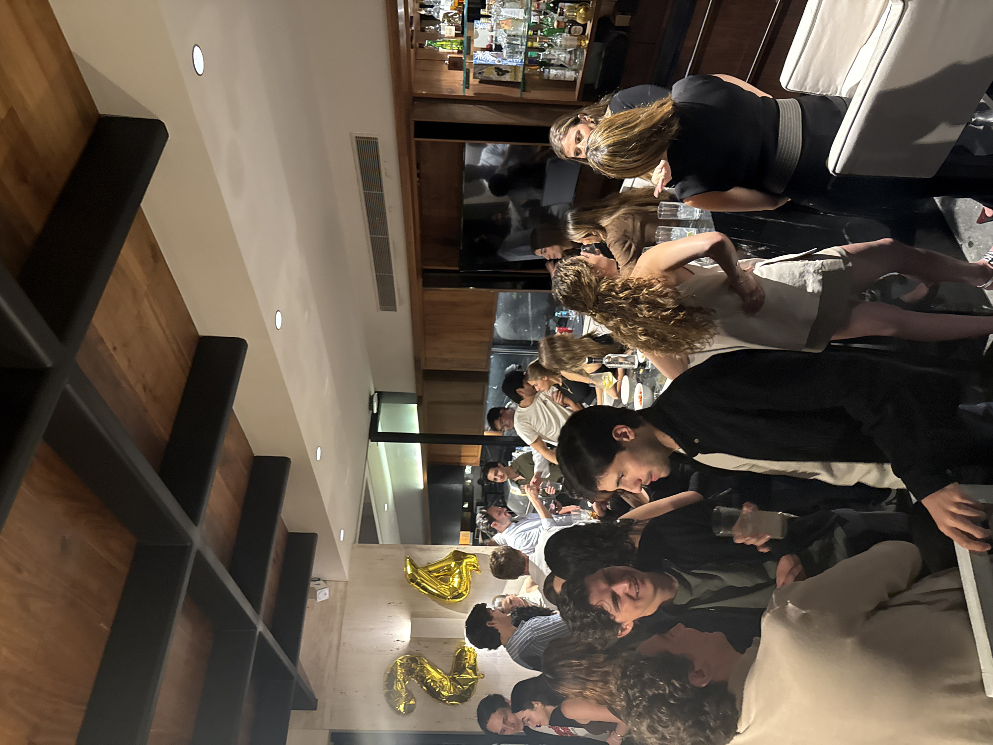 The scene depicts a gathering of people in what appears to be a restaurant or bar setting. The image shows a long table with numerous individuals seated and standing around it. Most of the people are interacting with each other, some holding drinks and engaging in conversation. The background reveals a well-stocked bar with various bottles. Gold-colored balloons in the shape of the number '2' are visible against a wall. The location is likely in Mexico City, Mexico.