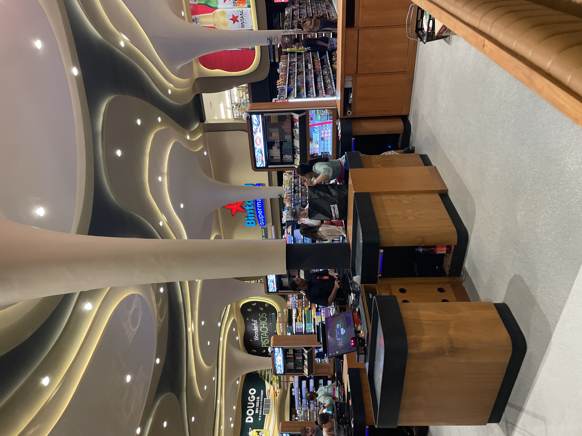 This image captures the interior of a modern supermarket or convenience store, focusing on the checkout area. A distinctive feature is the undulating, multi-layered ceiling, which incorporates numerous spotlights and indirect warm lighting, creating a sleek and contemporary aesthetic.
In the foreground and midground, several checkout counters are visible, constructed from light-colored wood with dark trim and integrated computer screens. Employees and customers are present; one person in a black shirt is behind a counter, and another individual wearing a light green top is at a checkout, seemingly processing purchases. Further back, additional shoppers can be seen browsing aisles stocked with various consumer goods.
Product displays are prominent, including a vertical refrigerated unit possibly holding beverages or tobacco products, and shelves filled with packaged items. A padded, light brown bench with a wooden frame is visible along the wall on the right side of the image.
Visible text includes branding like "DOUGO 100% CANOLA OIL IT'S YOUR HEALTH INVESTMENT", "Wonderful PISTACHIOS", and "BINTANG CRYSTAL". Store signage components such as "Binto superma" (likely part of "Bintang supermarket"), "ION", "Antang market", and "BIN" are also discernible. The setting suggests an indoor environment, making the time of day indeterminate.