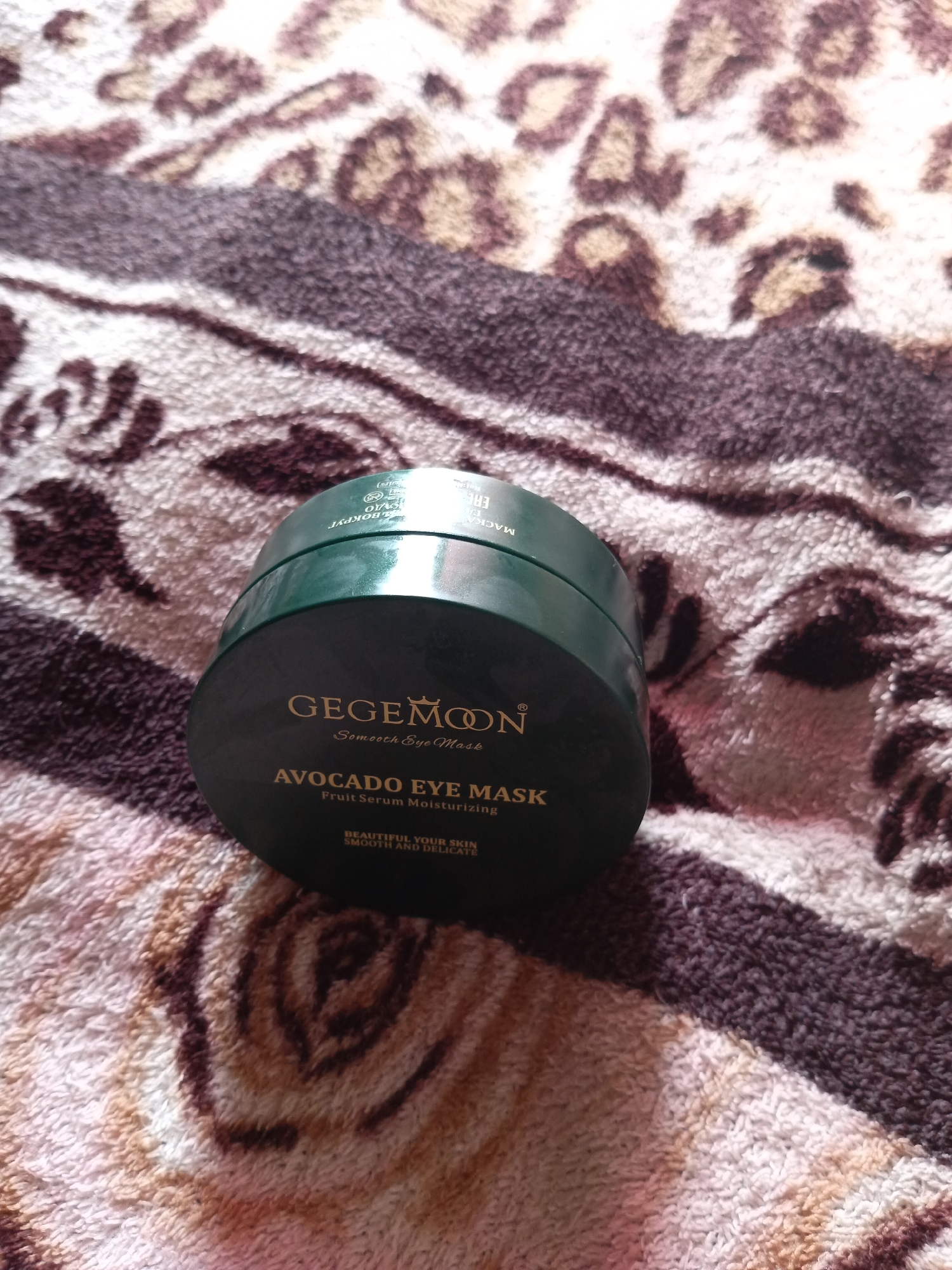 The image shows a dark green jar of GegeMoon Smooth Eye Mask with Avocado Eye Mask and Fruit Serum Moisturizing text printed on it. The jar is placed on a patterned fabric with leopard print details. The scene is indoors and the lighting suggests it is daytime. There are no people or animals visible in the image. The location is not specified as Monastirische, Russia; this information is not present in the image.
