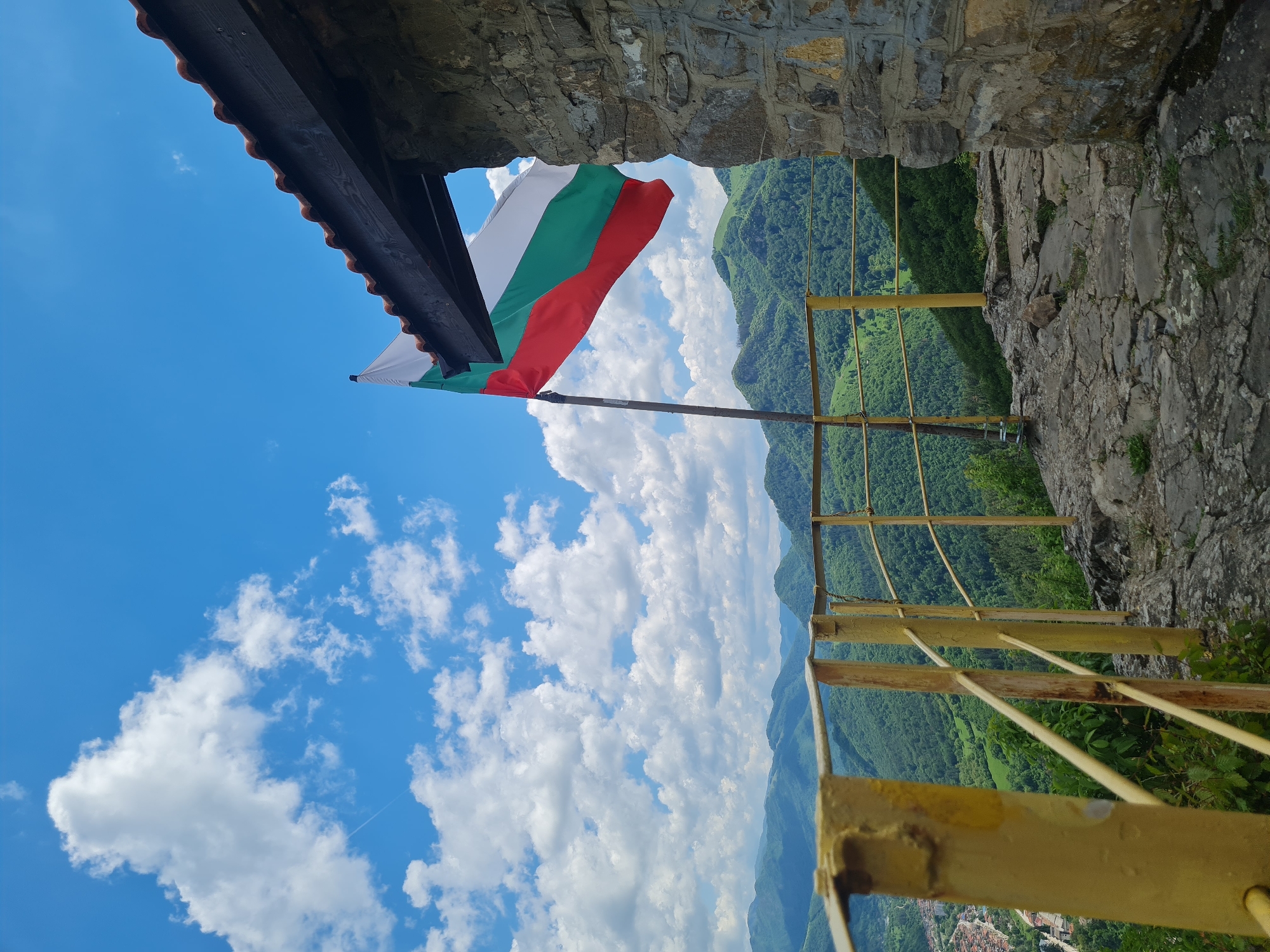 A ground-level, vertical, medium shot captures a view from a stone structure, likely a fortress or castle, with the Bulgarian flag waving against a backdrop of mountains, a clear blue sky, and scattered clouds.
The Bulgarian flag, featuring horizontal stripes of white, green, and red, is prominently displayed on a pole that is angled to the left. Part of the stone structure's roof is visible above the flag.
Beyond the stone structure, a mountainous landscape is visible, covered in lush green vegetation. The blue sky is dotted with white clouds, suggesting a clear, sunny day.
A yellow metal railing is positioned along the edge of the stone structure, possibly serving as a safety barrier for visitors. Below the mountains, a small town or settlement is faintly visible.