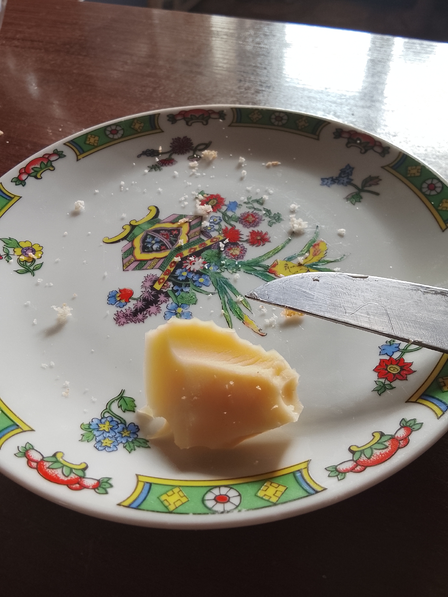 A close-up shot shows a piece of cheese on a decorative plate. The plate has a white base with colorful floral designs and geometric patterns along the rim. A silver knife rests on the plate, angled towards the cheese. Scattered crumbs are visible on the plate. The background appears to be a wooden table. There are no indications of the location being in Almaty, Kazakhstan.