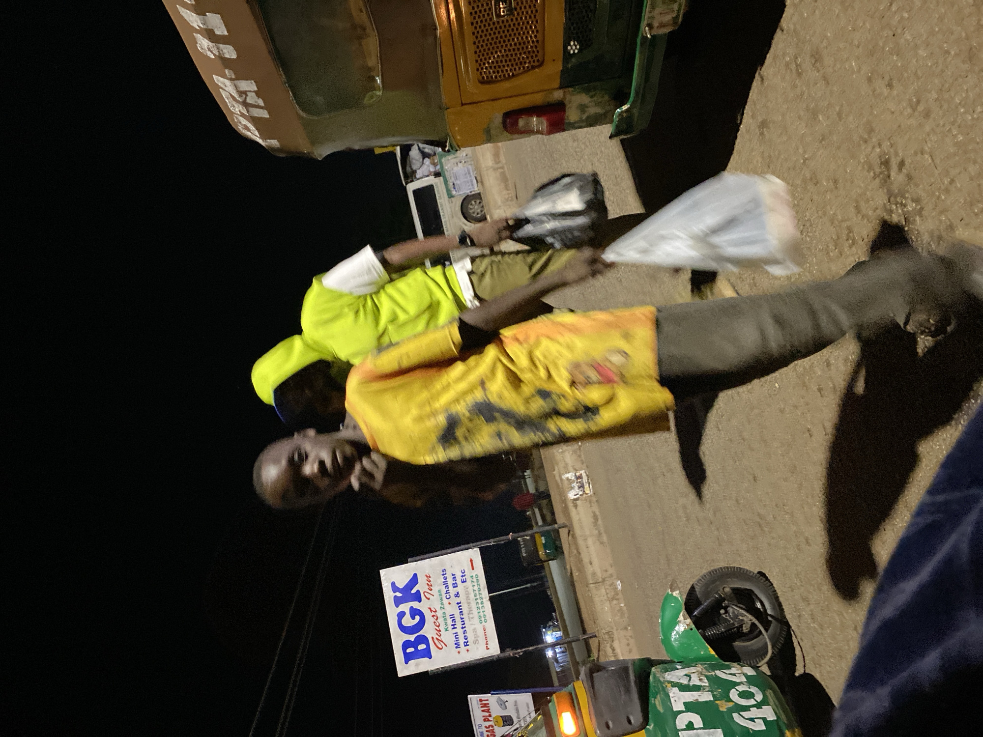In Bukuru, Nigeria, at night, a young person in a yellow t-shirt stands facing the camera, with their chin resting on their hand. Behind them, another person wearing a neon green hoodie and white t-shirt walks away, carrying a black plastic bag. To the left, a green motorized tricycle with "PTA 404" visible on its side is parked. A sign for "BGK Guest Inn" with details about its amenities and contact numbers is also present. In the background, a brown and green vehicle, possibly another tricycle, is visible. The scene appears to be a roadside in an urban area at night, with streetlights casting shadows.