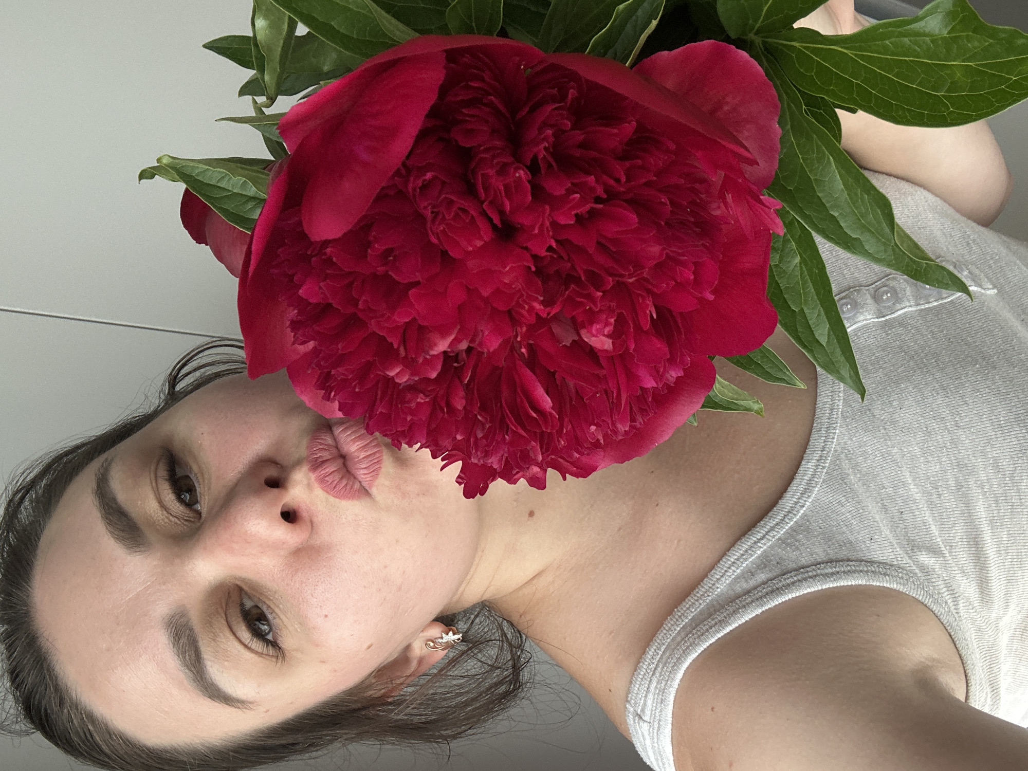 A close-up, rotated portrait captures a young woman playfully posing with a large, vibrant dark red peony flower. The woman, with dark hair and natural makeup, is visible from her upper chest to her forehead, positioned at a diagonal in the frame. She makes a "kissy" face with her lips pursed, looking directly towards the camera.
The voluminous peony, featuring richly textured, deep red petals and several broad green leaves, is held prominently next to her face, partially obscuring her right cheek. She is wearing a light gray ribbed tank top or camisole, with a small section of buttons visible on a placket near her shoulder. A small, star-shaped earring with a clear stone adorns her right ear.
The background is a plain, light gray or off-white wall, suggesting an indoor setting. The lighting is soft and even, creating a gentle and natural ambiance. The overall mood of the image is lighthearted and affectionate. There is no visible text in the image. The location context provided for this image is Chaikovsky, Russia.