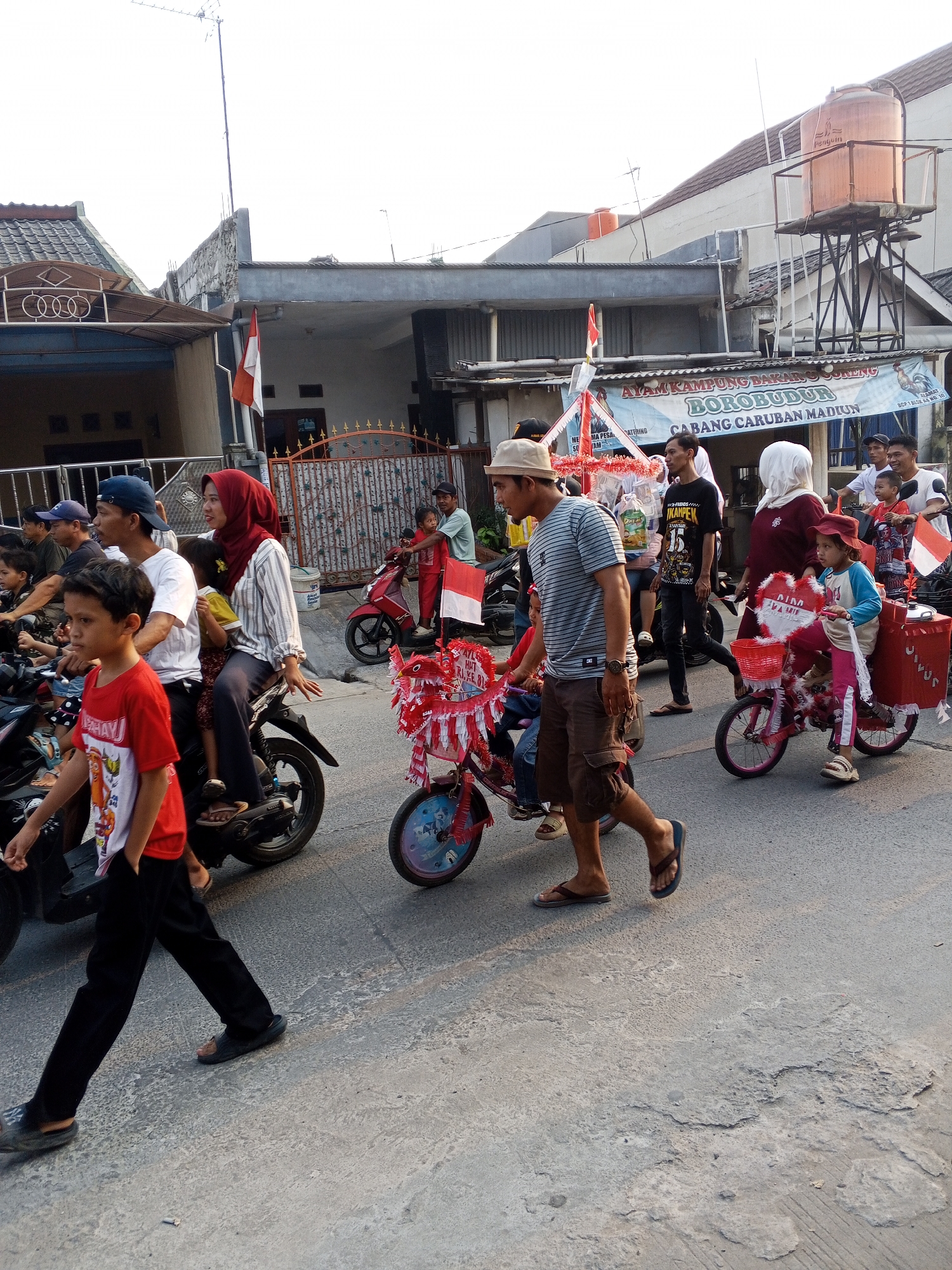 In Ciruas, Indonesia, an outdoor procession moves along a paved street. A group of people, including adults and children, are participating in the event. Some individuals are riding motorcycles, while others walk alongside. Several motorcycles are decorated with red and white streamers, suggesting a festive occasion. One child is riding a small pink bicycle adorned with similar decorations, and another child is seated on the back of a motorcycle holding a red and white flag. In the background, buildings line the street, and a water tower is visible. The sky is overcast.