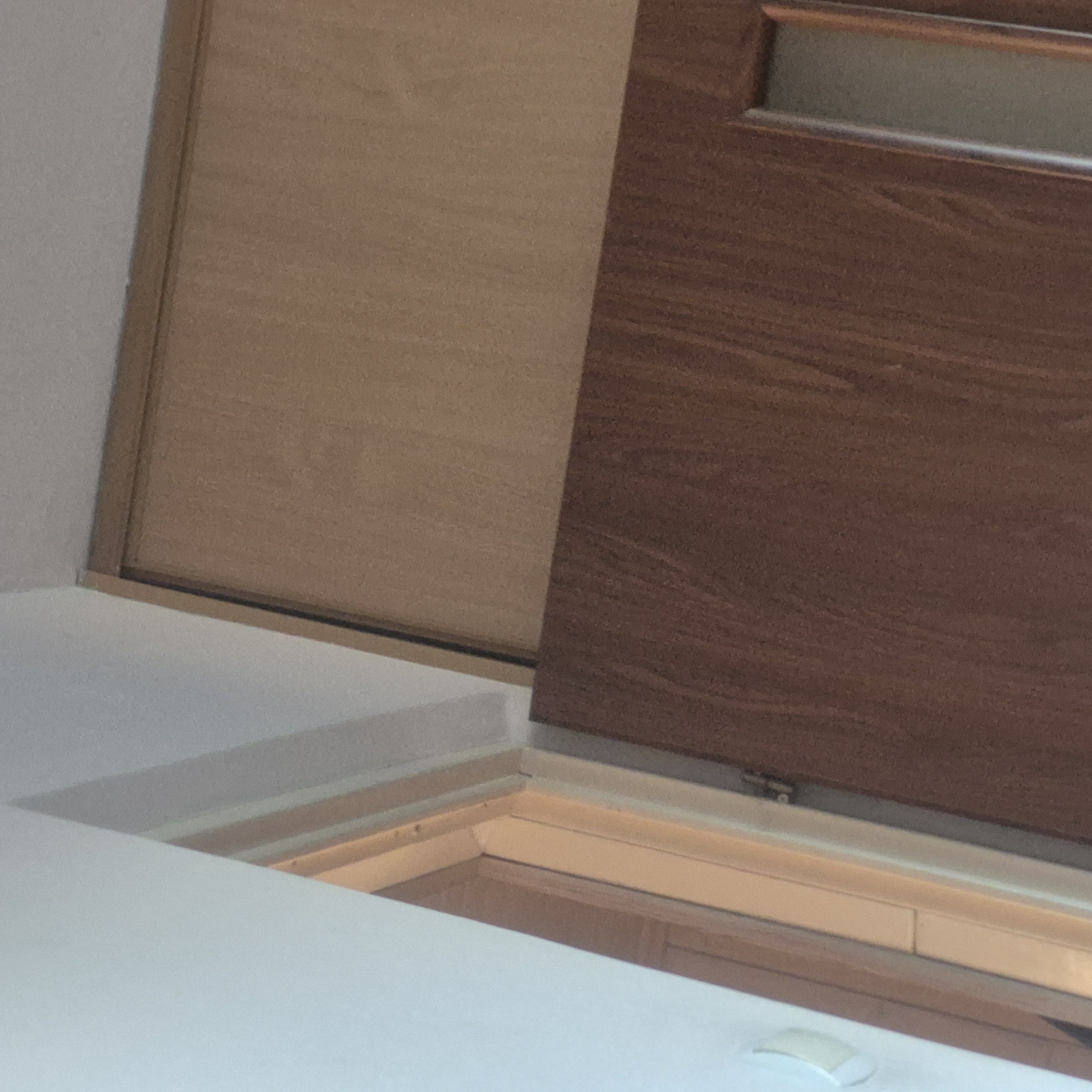 This is a close-up, interior, eye-level shot of a light-colored wall adjacent to a wooden door frame and a dark brown wooden door. The door is slightly ajar, revealing a sliver of another wooden door or partition on the left. The wall on the left is painted a light, slightly off-white color, and shows a slight sheen, suggesting it might be finished with satin paint. There is a slight shadow cast on the wall to the left of the door frame. The door frame is a lighter wood color than the door, with a slightly textured surface. The door itself is a darker brown with a prominent wood grain pattern, and it appears to be a solid door with a single vertical glass insert towards the right edge of the frame. The lighting is somewhat dim, with the light source appearing to be coming from the left side of the frame, casting subtle shadows. The scene appears to be a domestic interior, possibly a hallway or entrance to a room, and the image quality is somewhat grainy. There are no people or animals visible in the image. The scene is static and there are no discernible actions or interactions taking place. The location in the city of Kępno, Poland, is not discernible from the visual information in the image.
