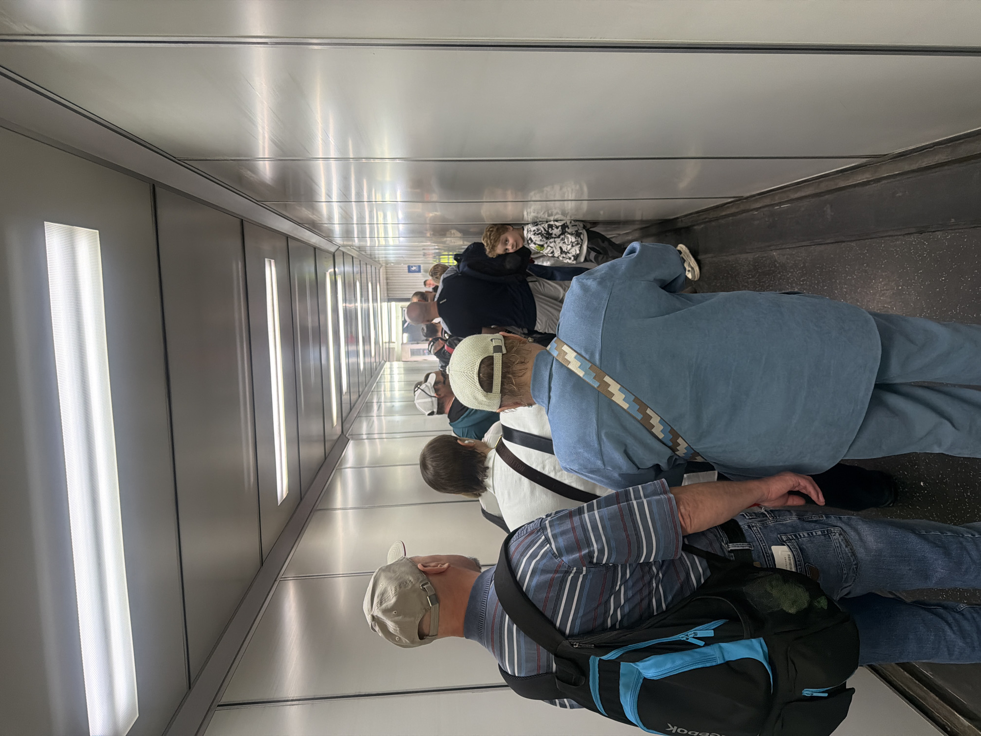 A photograph depicts a crowded pedestrian tunnel, possibly in Saint Petersburg, Russia. The tunnel is narrow, with light gray metal walls and ceiling. Vertical light fixtures illuminate the space. A group of people are walking through the tunnel, mostly facing away from the camera.
The individuals in the foreground include a man wearing a striped shirt, jeans, and a tan baseball cap, with a black and blue backpack. Behind him is a person with a white baseball cap and a white shirt with a dark strap. Next, there is a person wearing a blue jacket and blue pants. Further down the tunnel, there are other people. The tunnel appears to be a pedestrian walkway with a conveyor belt to assist in walking. The floor is dark gray with visible patterns. The overall scene shows a public space utilized by many individuals traveling in the same direction.