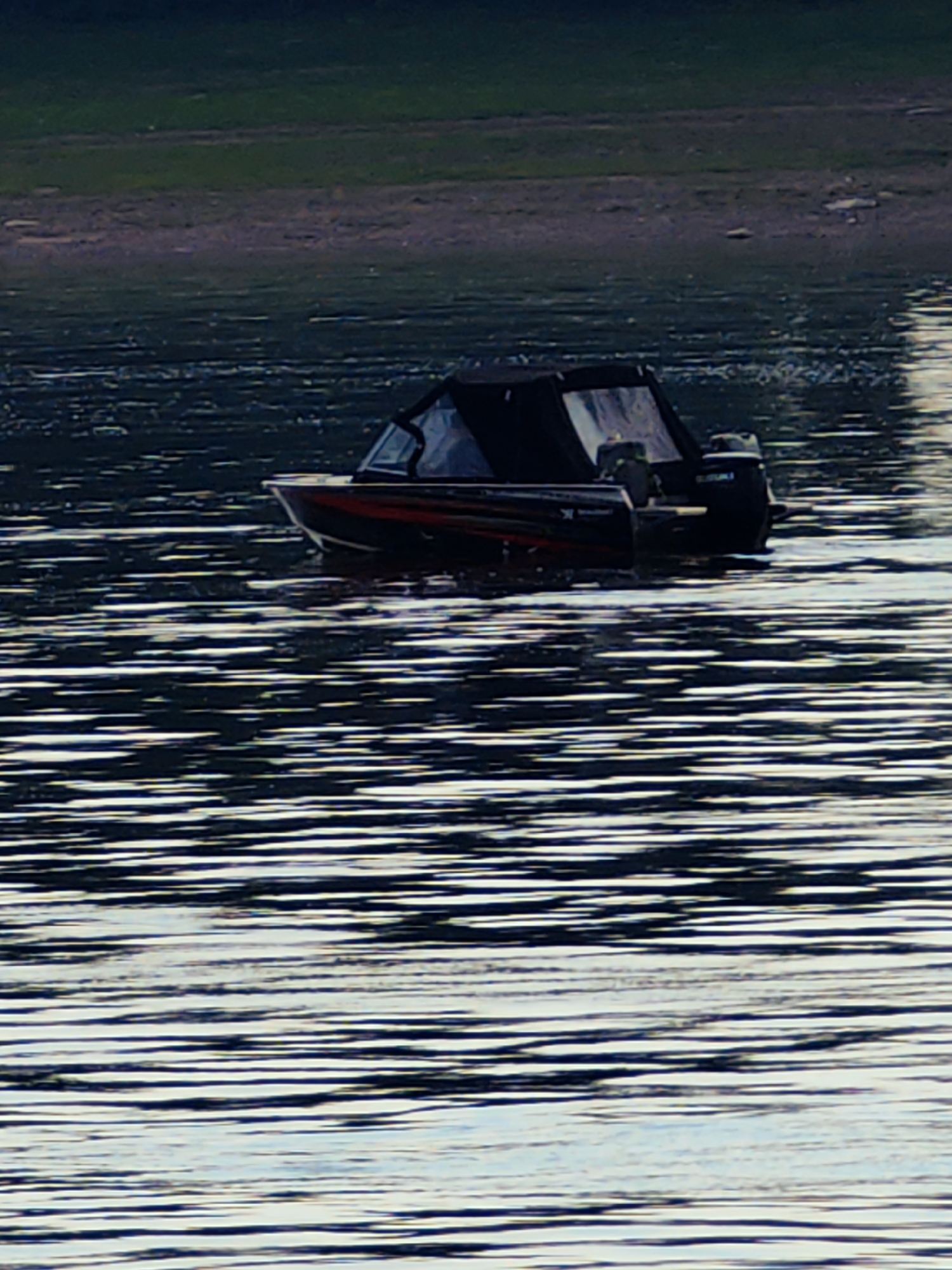 A black and red motorboat with a black canopy is moving on dark water, leaving a wake. The boat has a Suzuki outboard motor. The background shows a shoreline with green vegetation. The overall scene is likely a natural body of water, possibly a lake or river, and the image does not provide any specific information about the city of Erkalovo, Russia.