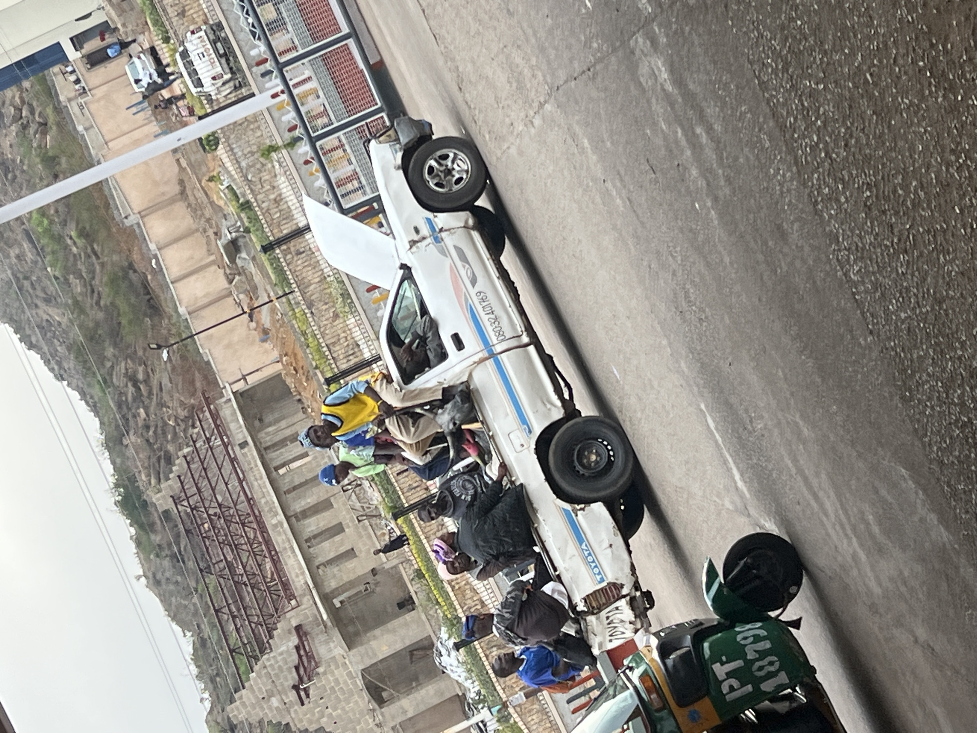 The image captures an outdoor scene in Jos, Nigeria, depicting a bustling area with people and vehicles. The perspective is slightly elevated, looking across a road or open space towards a hillside and construction sites.
The primary subjects are a white pickup truck, identifiable as a Toyota, and numerous individuals clustered around it. The truck appears to be a commercial or shared transport vehicle, as evidenced by the group of people, many of whom are men, standing near and inside it. Some individuals are wearing casual attire, including a man in a yellow vest and others in caps. One person is visible inside the cab of the truck. The truck shows some wear and damage, particularly on its front passenger side. On its side, the number "08032401769" is displayed, possibly a contact number, and the brand "TOYOTA" is clearly visible on the front.
In the immediate foreground, partially visible on the right, is a green and yellow motorized rickshaw, commonly known as a Keke Napep, with "PT-" and "18798" visible on its front.
The background features a rocky, uneven hillside on the left, suggesting natural terrain. In the mid-ground, there are multiple unfinished buildings under construction, characterized by exposed concrete blocks and metal framework for roofing, indicating ongoing development. Fences or barriers are also visible in this area. The sky appears bright but overcast or hazy, suggesting daytime. No strong emotions are discernible from the people; they mostly appear neutral or engaged in observation. The scene portrays a typical local transport hub or stop.