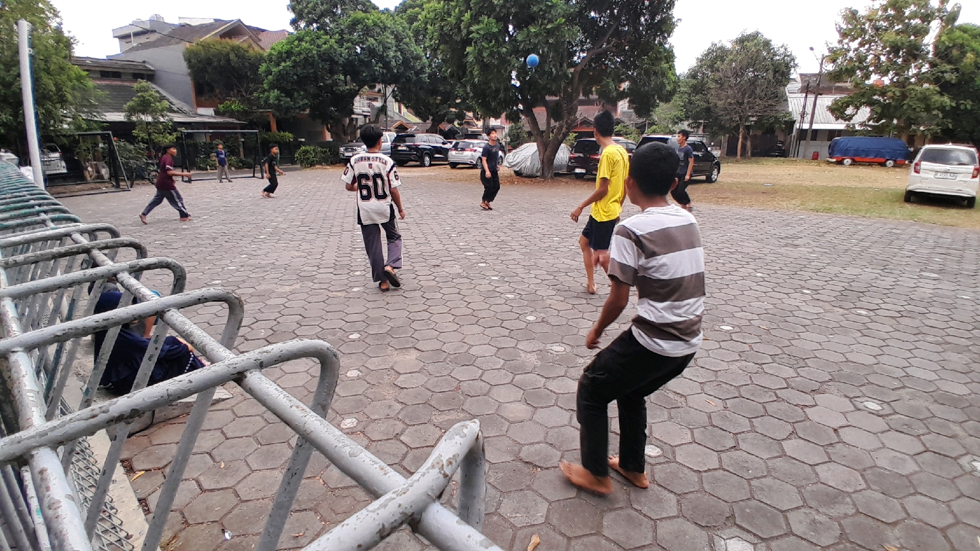 In a paved outdoor area with hexagonal tiles, several young males are engaged in a game of soccer. The scene is set in what appears to be a park or a recreational space, with trees and buildings visible in the background. One individual, wearing a white and black jersey with the number 60, is positioned mid-field, facing away from the camera. To his right, a person in a yellow t-shirt and dark shorts is also facing away, appearing to be actively playing. In the foreground, another young person in a striped t-shirt and black pants is captured in motion, with one leg extended as if kicking or dribbling a ball, though the ball itself is not clearly visible in this shot. Further back, a person in a maroon shirt and dark pants is walking away from the camera, and another individual in black is running. A blue ball is in mid-air near the trees. A silver car is parked in the background on the right side of the frame. On the left, metal fencing surrounds a seating area where a person wearing a dark hijab is seated. The overall atmosphere suggests a casual, outdoor activity in a moderately urban or suburban environment in Bandung City, Indonesia.