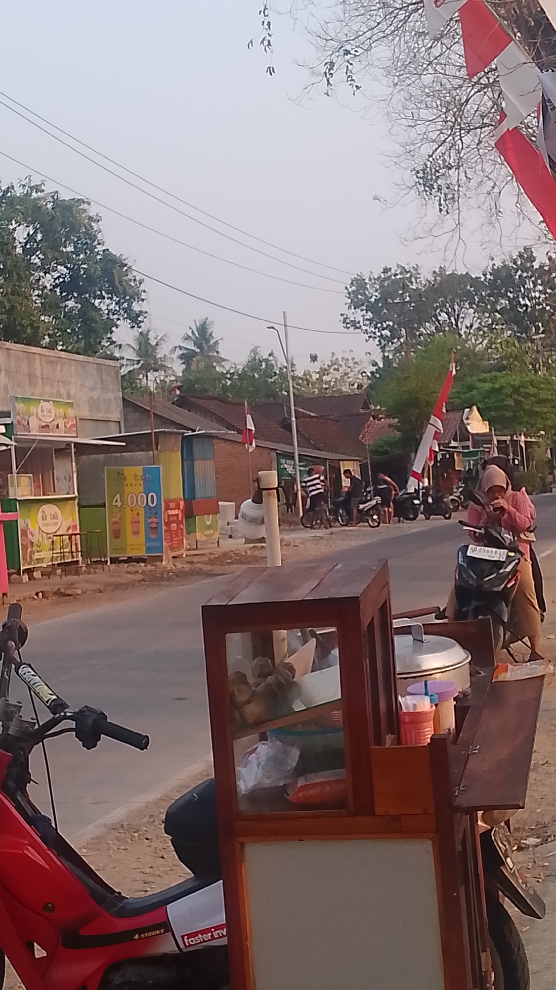 The image depicts a street scene in Karanglangu, Indonesia, likely during daylight hours given the lighting. In the foreground, a red motorcycle is parked, with its handlebars and front section visible. Adjacent to the motorcycle is a wooden food cart with glass display cases. Inside the cart, various food items and containers are visible, including what appears to be a large metal pot on top.
The midground of the image shows a road with several motorcycles parked and a few individuals. One woman wearing a hijab and a pink top is seated on a black motorcycle, holding a cup to her mouth. Another person is standing next to her. Further down the road, several other motorcycles are parked, and a few people are standing or interacting with the parked vehicles.
The background reveals a street lined with buildings, some appearing to be shops or stalls. A prominent sign with the number "4.000" and images of drinks is visible on one of the stalls. Red and white Indonesian flags are displayed on poles in front of some of the buildings, indicating a local celebration or patriotic display. Trees and power lines are also present, contributing to the urban and semi-rural environment. The sky is overcast or hazy.