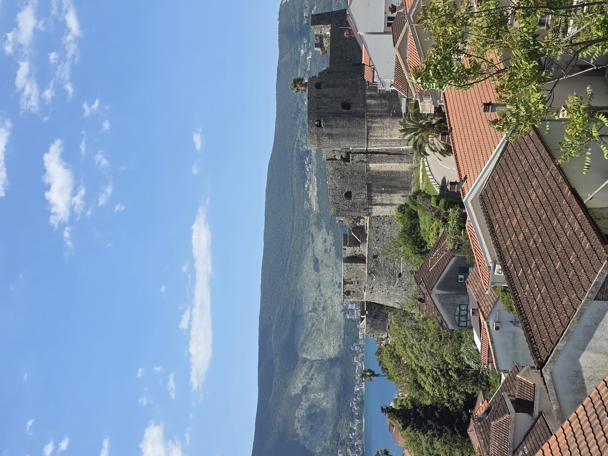 The image shows a view of Herceg Novi, Montenegro. An ancient stone wall runs vertically through the center of the frame, adjacent to a road. A mountain covered with green vegetation rises behind the wall. To the right of the wall, there are houses with brown tiled roofs. Some trees are visible among the houses. In the background, there is a blue body of water. The sky is blue with some white clouds. There are no people visible in the image.
