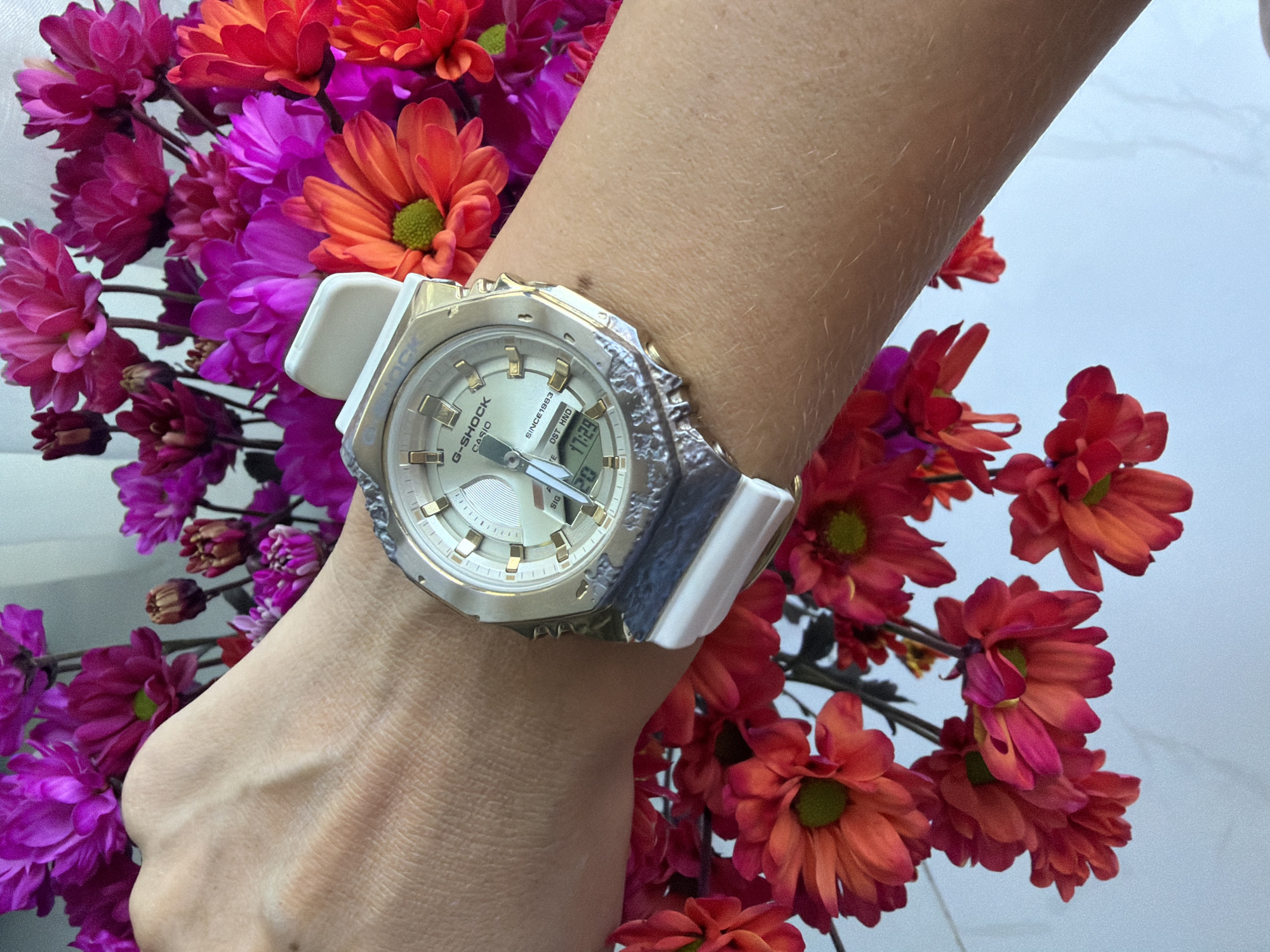 A close-up shot features a person's arm wearing a white and silver G-Shock watch. The watch face is a mix of analog and digital displays, with the time showing 11:29 and "DST HND" visible. The watch has a distressed silver bezel and white straps. The arm is positioned among a vibrant arrangement of artificial chrysanthemums in shades of magenta, fuchsia, and orange. The background is a pale, textured surface, possibly a table or wall, with a hint of light blue suggesting natural light. The overall composition is bright and colorful, with the focus on the watch and the flowers. There are no discernible weather cues or specific location cues beyond the flowers which may be typical of Nha Trang Ward, Vietnam. No specific emotions are conveyed.