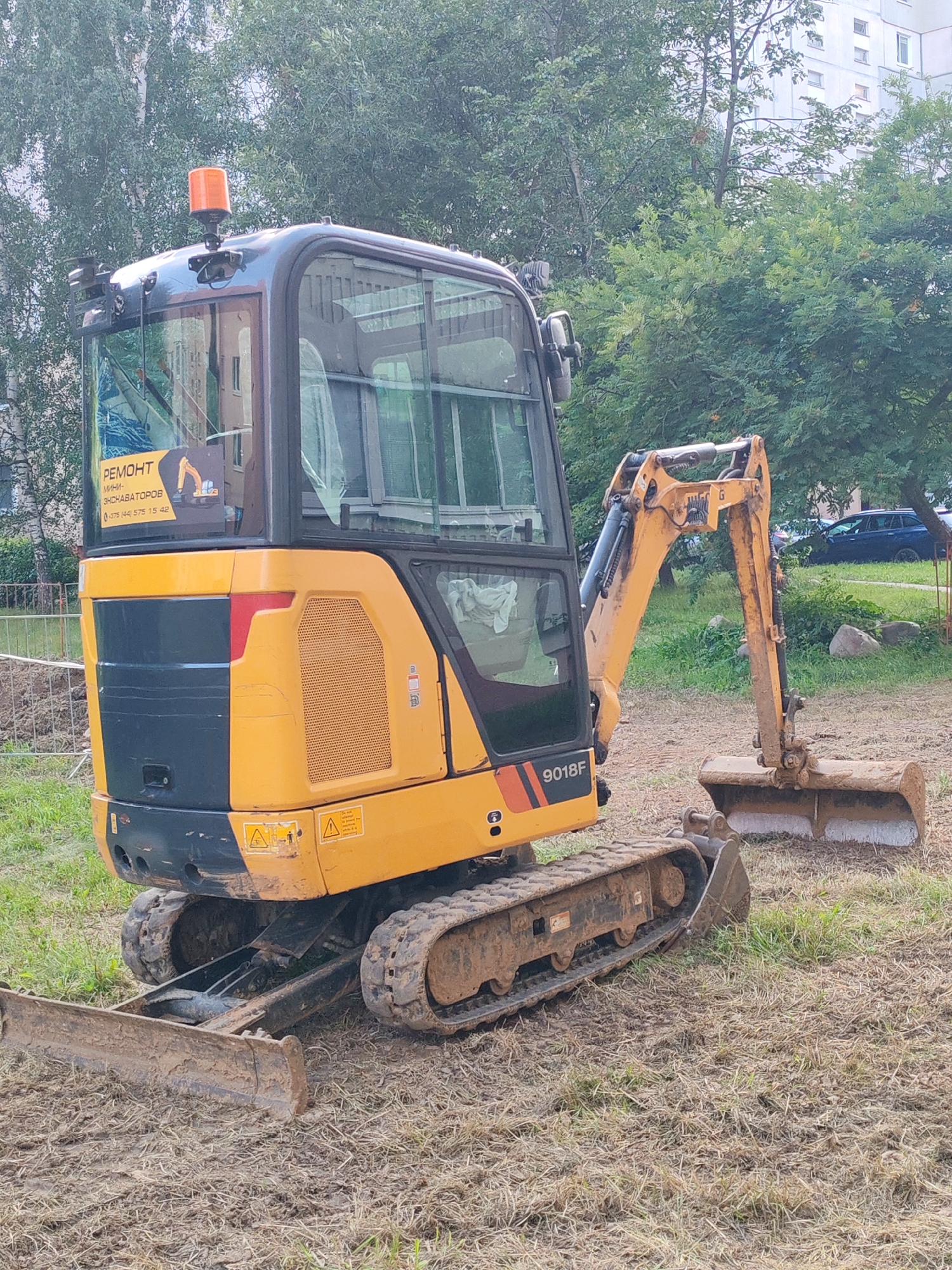 A yellow mini-excavator is parked on a patch of dry, patchy grass and dirt. The excavator is positioned facing right, with its tracks resting on the ground. The machine has a black and yellow body, a glass-enclosed cab, and an orange rotating beacon light on top. A sticker inside the cab window reads "РЕМОНТ МИНИ-ЭКСКАВАТОРОВ" and a phone number. The background features green trees and a blurred view of apartment buildings. A blue car is visible in the distance to the right of the excavator. The overall scene appears to be an outdoor, urban environment during daylight.