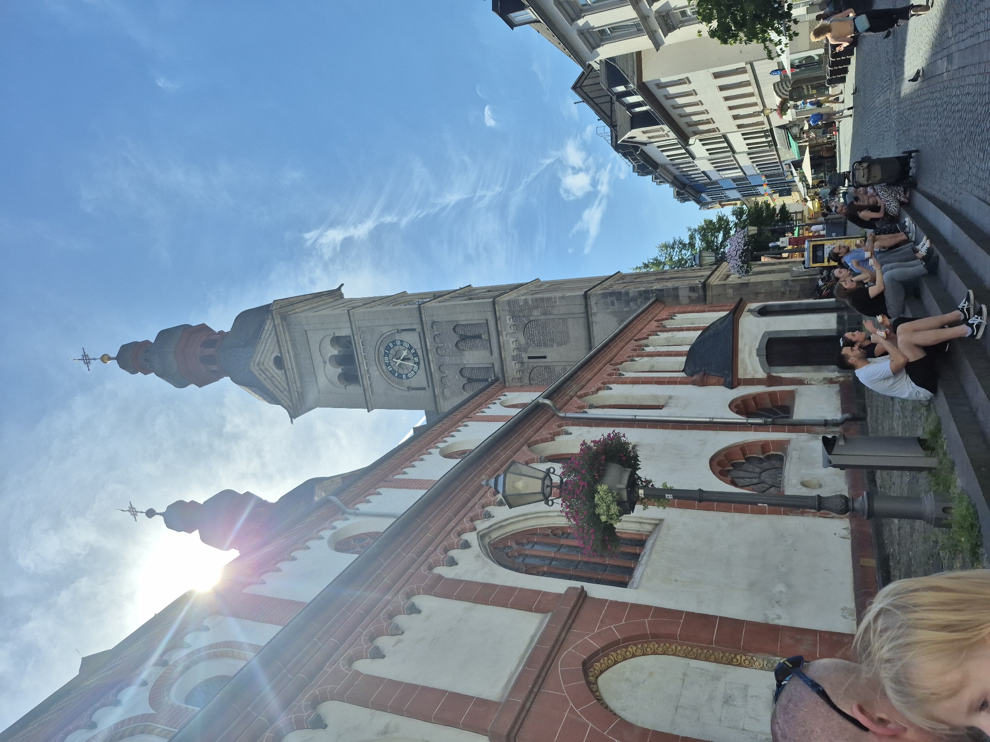 This is a low-angle, wide shot of a street scene in Koblenz, Germany, featuring the St. Castor's Basilica. The sun is bright and high in the sky, with some wispy clouds. In the foreground, a portion of a person's head and shoulder is visible, looking towards the left. To their right, another person's blonde hair is visible. The main subjects are the facade of the St. Castor's Basilica, a tall clock tower attached to it, and a street lined with buildings.
The Basilica's facade is white with red trim and arched windows. A street lamp with hanging flowers stands in front of it. The clock tower is made of stone, with a clock face visible showing approximately 1:08. People are sitting on the steps leading up to the Basilica, some eating ice cream. Further down the street, more people are walking and sitting. The street is paved with cobblestones. The buildings lining the street are of a traditional European architectural style, with multiple stories and visible windows. The scene conveys a sense of a lively, sunny day in a historic European city.