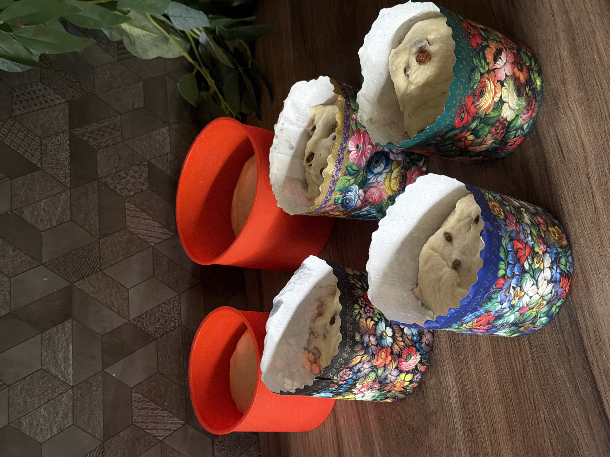 The image shows four cylindrical baking molds, two orange and two decorated with floral patterns. All four molds are lined with white paper and contain dough. The orange molds are empty, while the floral molds contain dough with what appear to be raisins. The molds are arranged on a wooden surface that is partially overlaid with a darker, patterned surface. Green foliage is visible in the upper left corner of the image. The location is not specified, and there is no specific indication that the location is in the city of Perm, Russia. There are no people in the image.