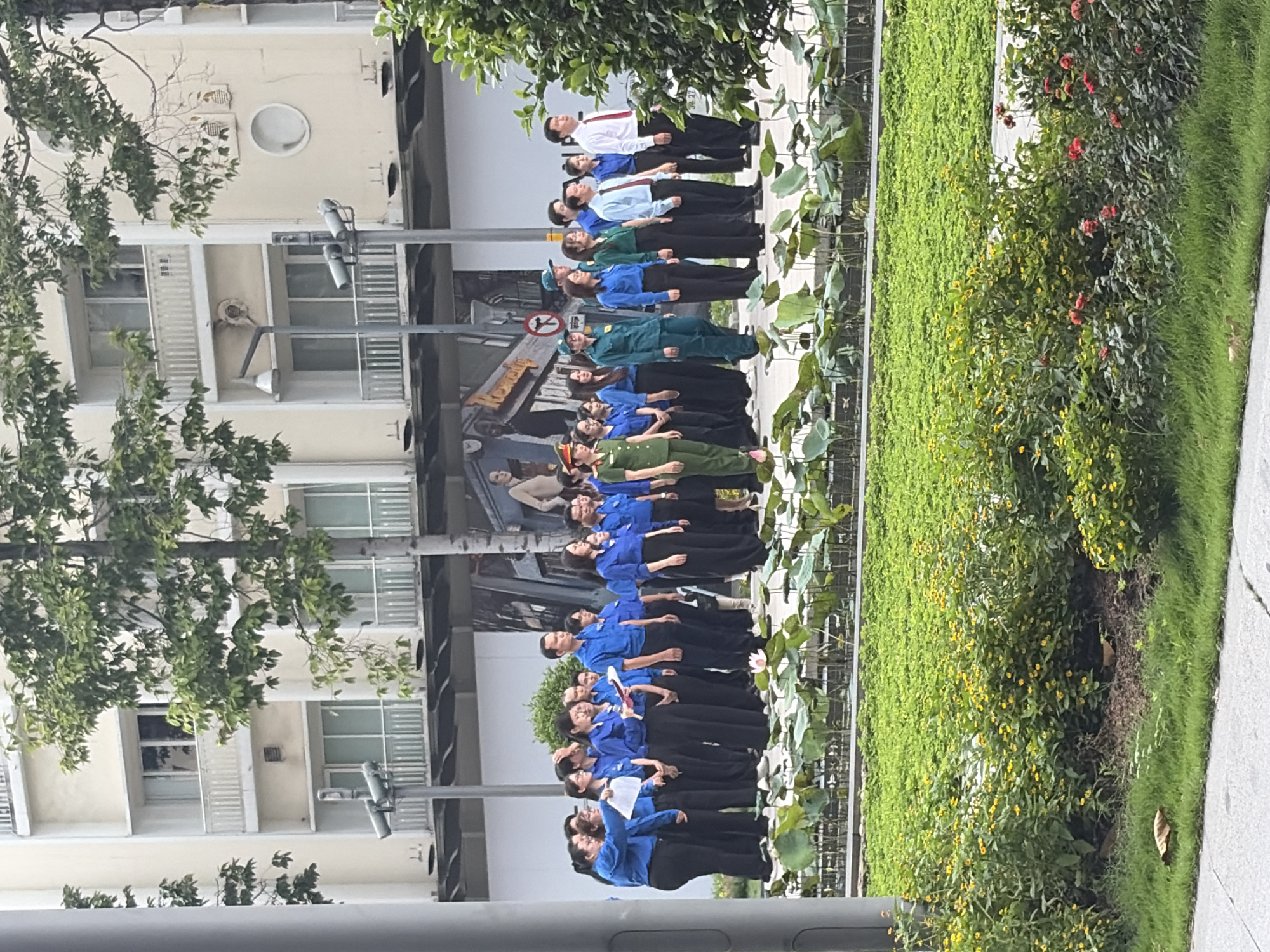 An outdoor scene in Thủ Đức, Vietnam, shows a group of approximately 25-30 individuals standing in a loose formation, appearing to be lined up for an event or gathering. The majority of the group, primarily women, are dressed in matching blue long-sleeved shirts and black trousers, with several holding papers or small books. One individual in the group wears a green military-style uniform with epaulets and a cap. Another man at the far right wears a white collared shirt with a red tie and dark trousers, while other men in the group wear blue shirts similar to the women.
In the immediate foreground, a meticulously maintained urban garden features a strip of green grass, several bushes with small yellow flowers, and a section with red roses. Beyond this, a shallow water feature contains numerous large, round green lily pads (lotus leaves), with a single pink lotus flower in bloom on the left side of the feature.
Behind the people, a paved area separates them from the background structures. The background consists of multi-story buildings with light-colored facades, featuring numerous windows and balconies with white railings. Large leafy trees are interspersed between the buildings and the paved area. Street furniture, including a grey pole with a lamppost and two surveillance cameras, is visible. A large mural or advertisement depicting human figures covers a section of a building wall behind the group. A red circular "no turn" or "no entry" traffic sign is partially visible on a pole behind the group.
