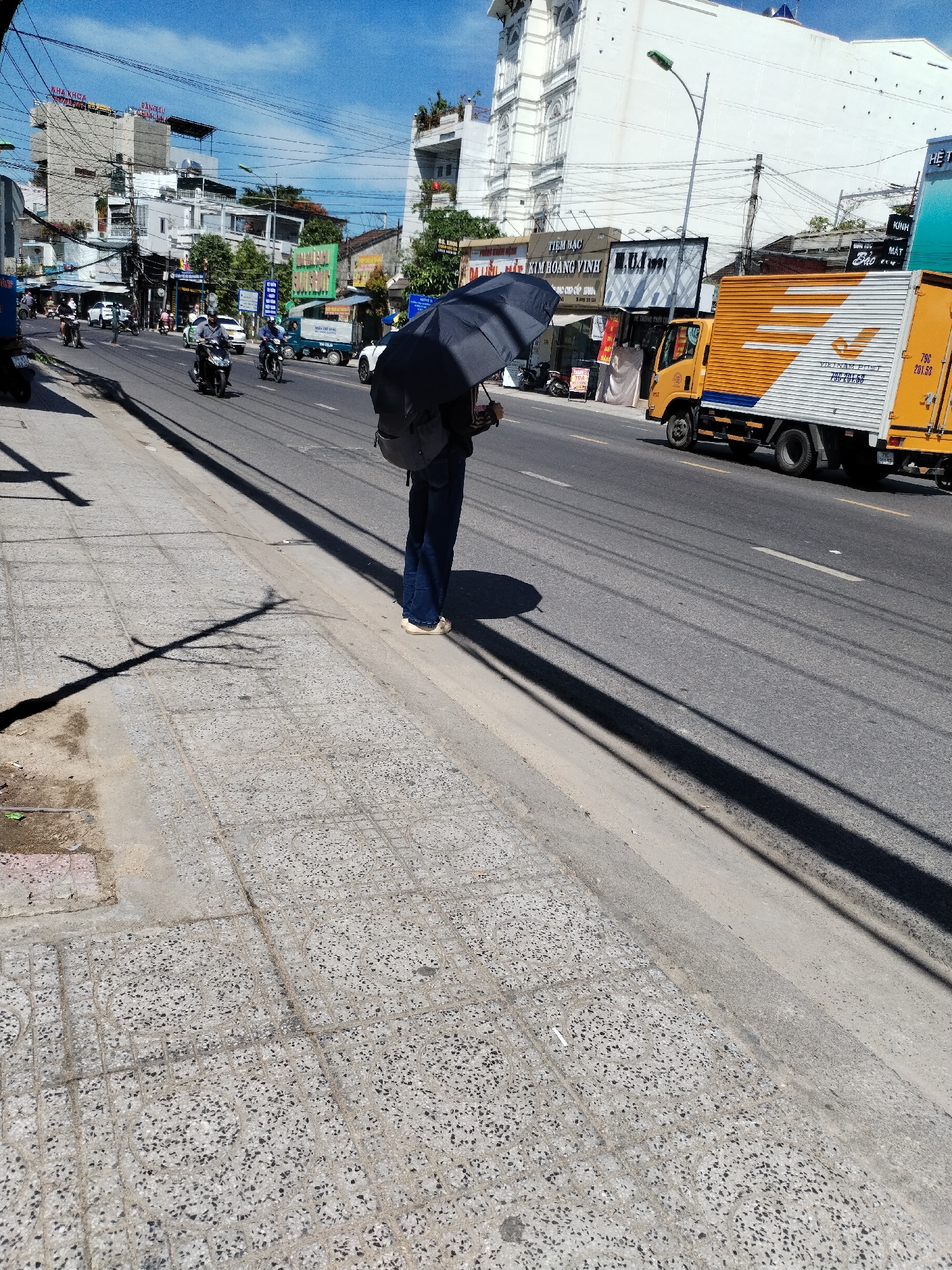 A lone individual stands on a sidewalk next to a busy street in Nha Trang, Vietnam. The person is holding a dark umbrella open, casting a long shadow across the textured concrete sidewalk. They are wearing dark pants and a backpack. The street is filled with traffic, including motorcycles, cars, and a yellow delivery truck with "VIETNAM POST" written on its side. Buildings line the street, some with signage in Vietnamese. The sky is clear and blue. The scene depicts a typical day in a bustling Vietnamese city.