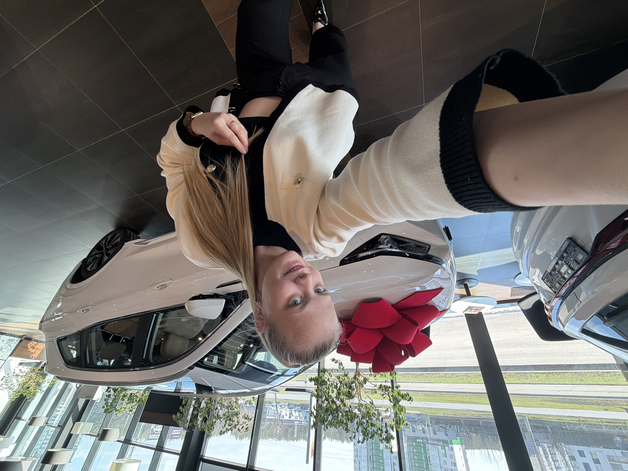 A selfie-style shot captures a young woman inside a modern car dealership, looking directly at the camera with a slight smile. She has light-colored hair and is wearing a cream-colored cardigan with black trim and gold buttons over a black top. Her left hand, adorned with a ring and a bracelet, is visible near her shoulder.
The setting appears to be a new car handover, with a white SUV or crossover prominently featured in front of her, decorated with a large red bow on its hood. Another white vehicle is partially visible to the right. The dealership has a dark, tiled floor and expansive windows that offer a view of the outside world.
Beyond the windows, an urban scene unfolds, featuring a multi-lane road, trees, and several buildings under clear daylight conditions. Hanging plants add greenery to the dealership's interior decor. Visible text on a license plate frame reads "BAULITE EXCLUSIVE" and "AVTOMAX". On a distant building outside, Cyrillic text "АВТОДОМ" (Avtodom, meaning Car Home) and "RENT" are discernible, consistent with the Izhevsk, Russia location. The overall atmosphere suggests excitement and a significant purchase event.