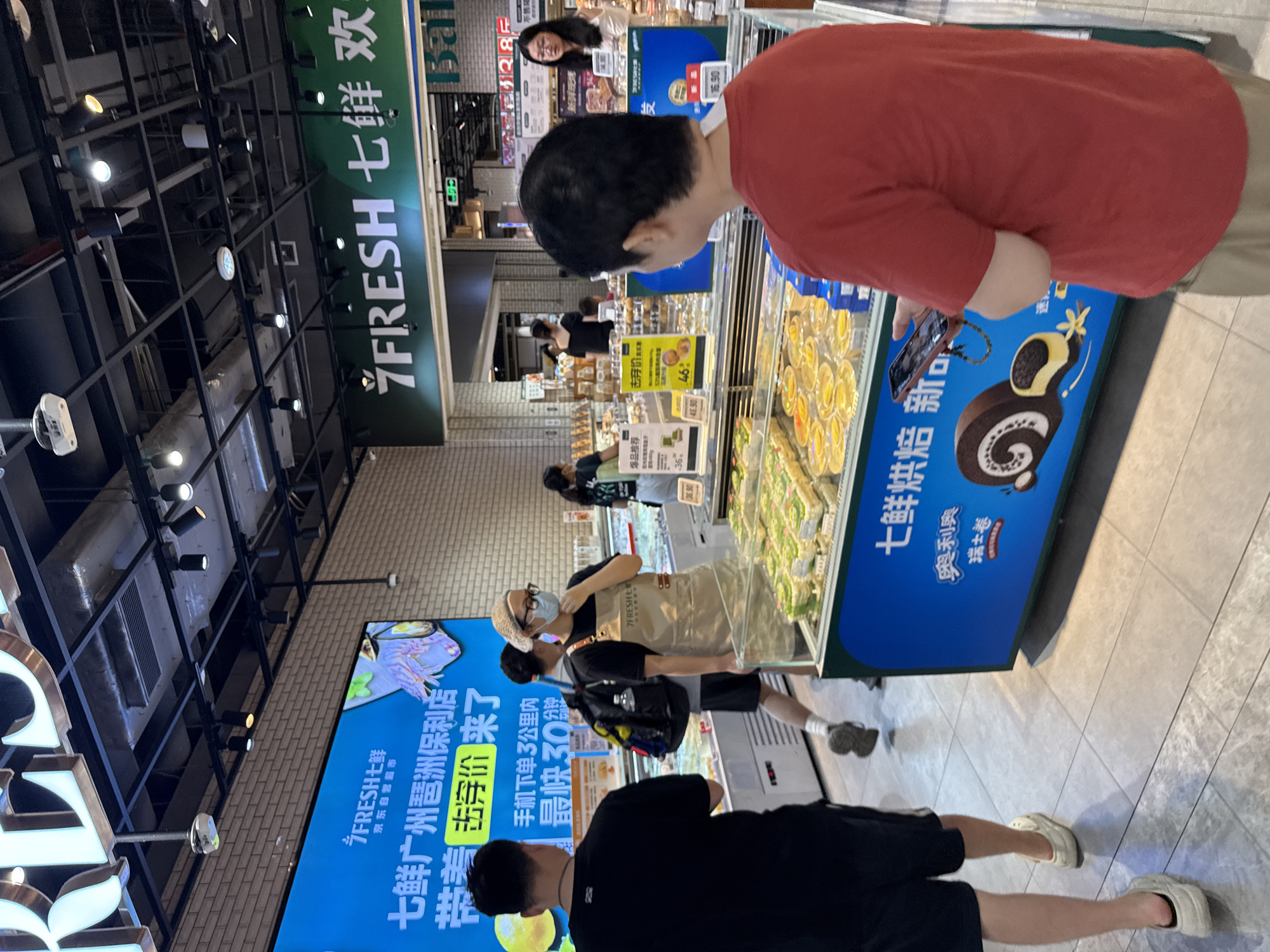 The image shows the interior of a 7FRESH supermarket in Haizhu District, China. In the foreground, a person in a red t-shirt is holding a smartphone. Behind them, several other shoppers are visible, including a woman in an apron wearing a face mask. Display cases filled with pastries and desserts are arranged throughout the store. A large digital billboard displaying promotional information is visible on the left side of the image, and a "7FRESH" sign hangs above a green banner on the right. The environment appears to be a well-lit, modern retail space with tiled floors and a suspended ceiling.