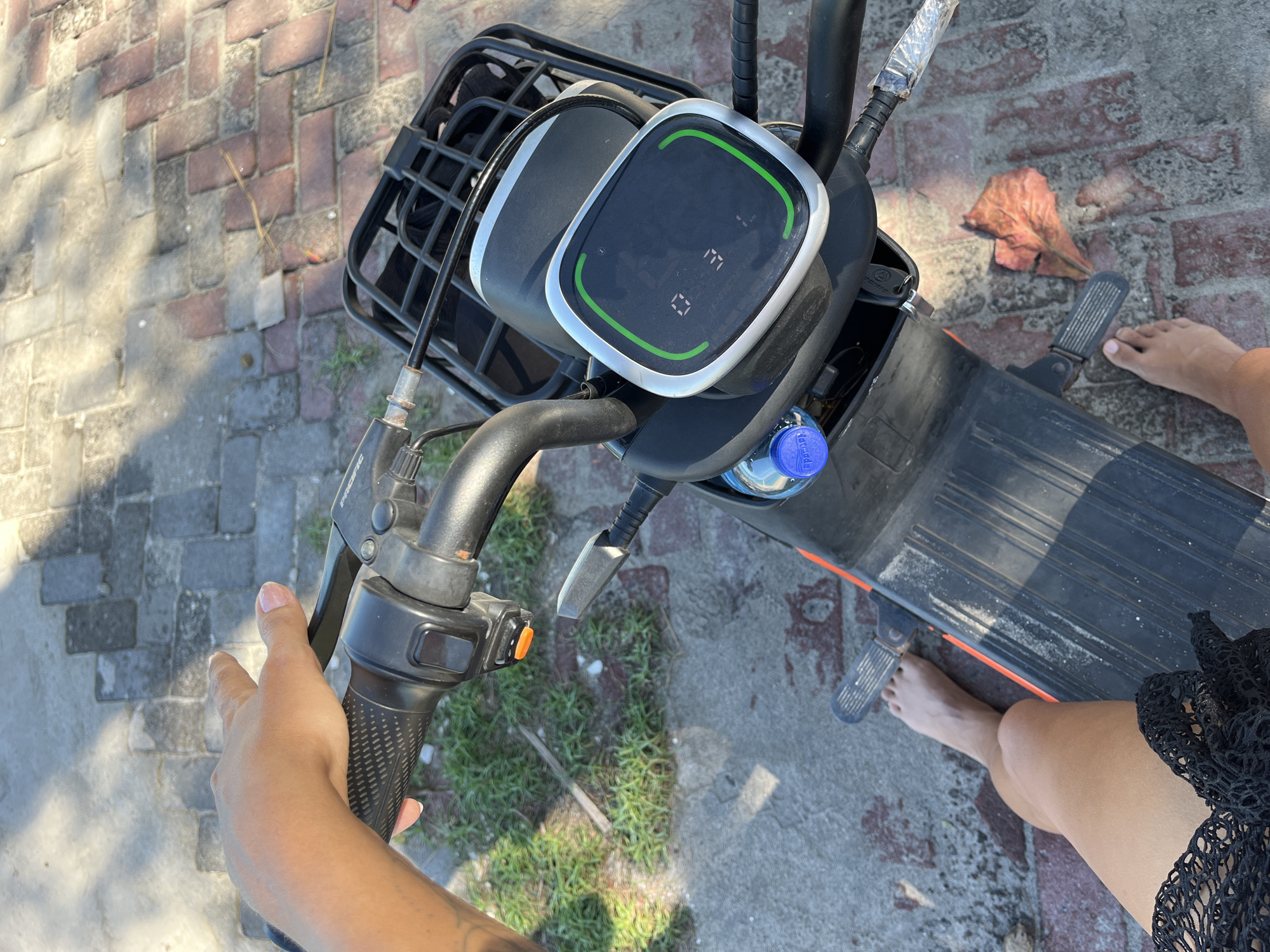 A first-person perspective photo shows the handlebars of a black electric scooter, held by a tan-skinned hand with a visible tattoo. The scooter has a front basket and a digital display with a green border. A blue water bottle is positioned near the display. The scooter is parked on a paved area with brick and concrete sections. Bare feet and legs are visible, suggesting the rider is wearing a black lace garment. A fallen leaf rests on the ground near the scooter's footrest. The location appears to be an urban area in Indonesia.