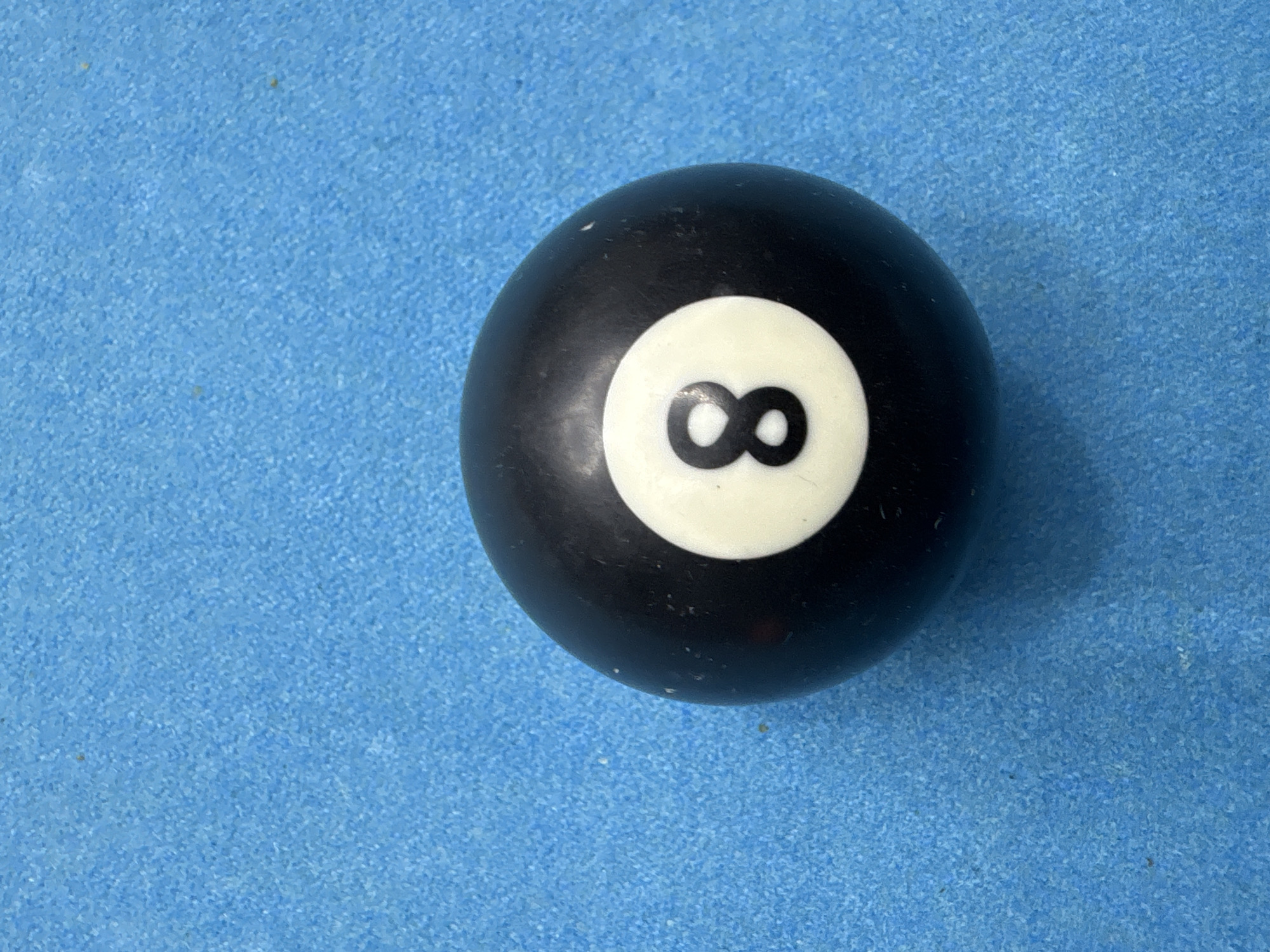 A close-up, top-down view shows an 8-ball resting on a light blue, textured surface. The 8-ball is predominantly black, with a white circular area displaying the black number "8". No people, actions, interactions, or other objects are visible. The location, Baan Bang Rak, Thailand, is not identifiable from the visual information.