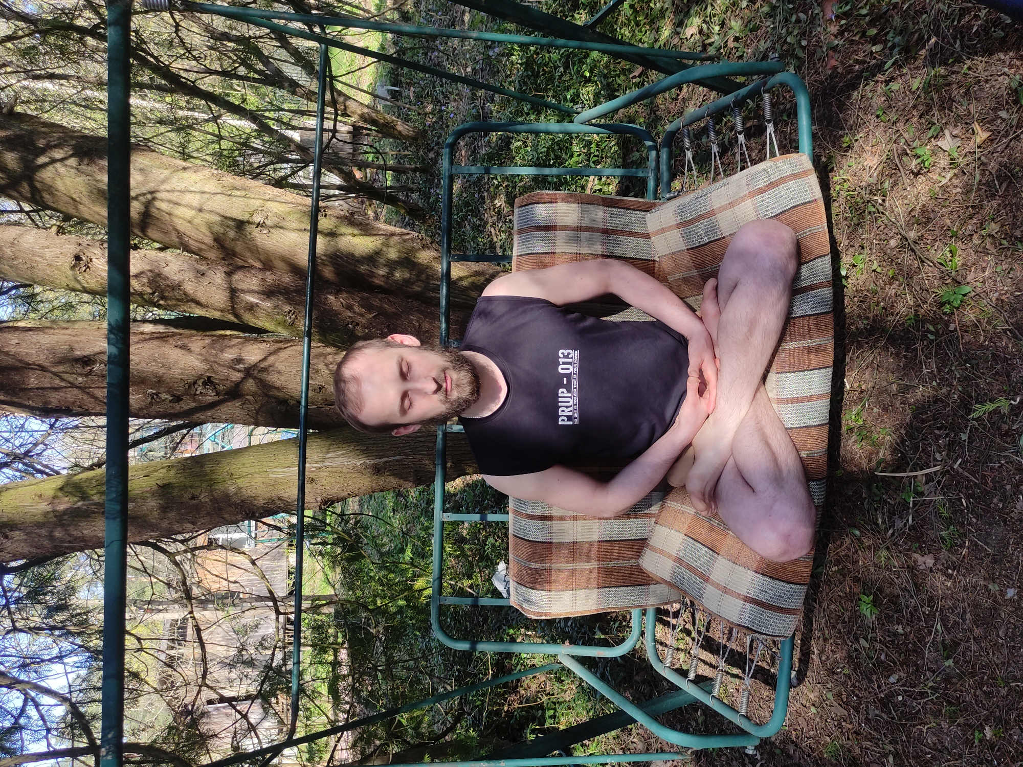 A man with a beard is sitting in a meditative pose on a swing seat outdoors. He is wearing a black tank top. The swing is made of green metal tubing and has a striped brown, beige, and green cushion. The background shows a tree and foliage. The location is not identifiable as Kratovo, Russia from the image provided.