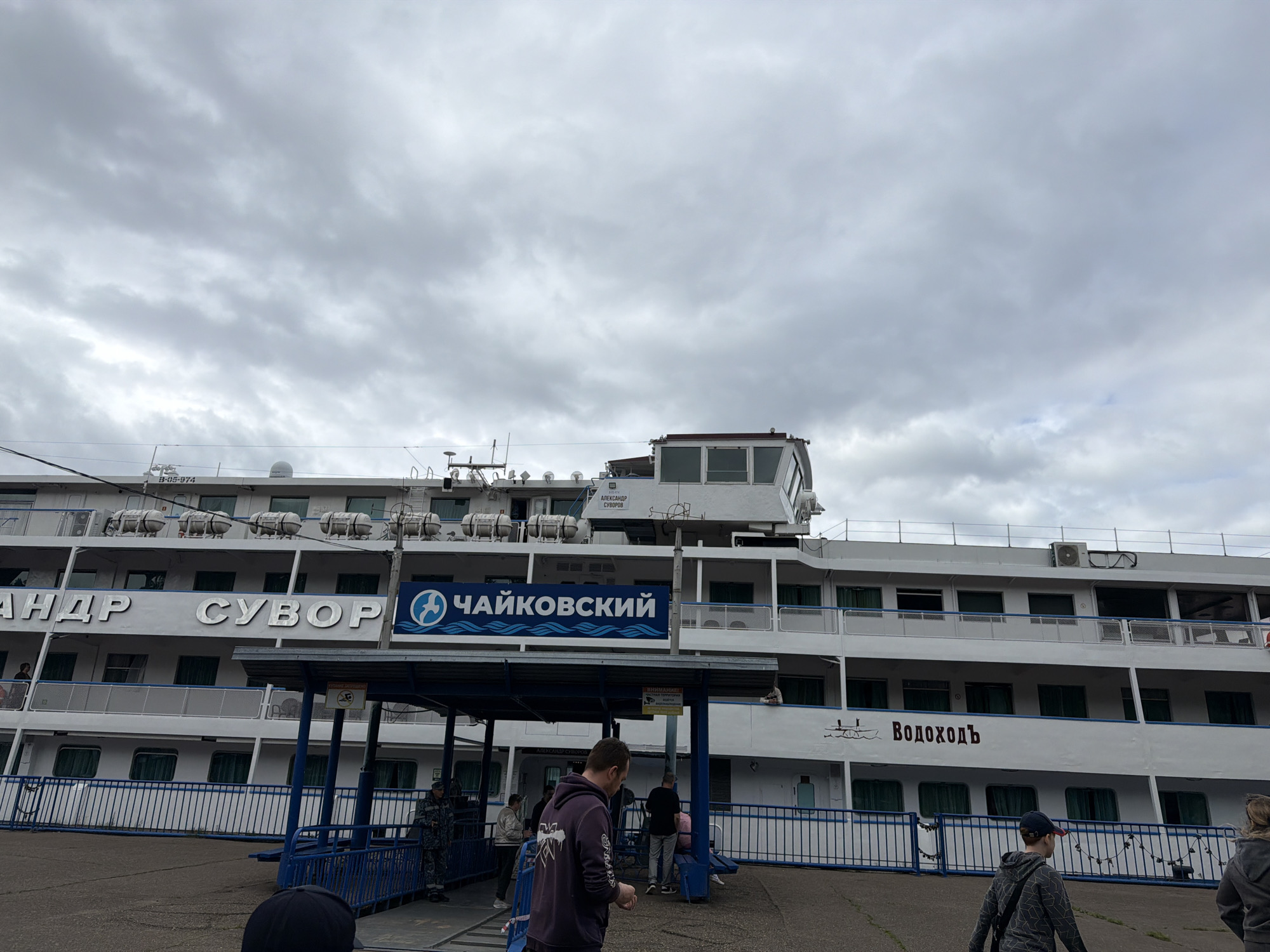 A wide shot captures a passenger ship docked at a pier under a cloudy sky. The ship, a multi-deck vessel, has "АЛЕКСАНДР СУВОРОВ" in large silver letters on its side, and further along, "ВОДОХОДЪ" in dark red letters. Above the pier, a blue sign with a circular logo and the text "ЧАЙКОВСКИЙ" is prominently displayed. Several people are visible on the pier and boarding the ship. One person, wearing a dark purple hoodie, stands looking down. Others, including a person in a dark uniform and a few individuals in casual clothing, are in the background near the entrance to the ship. The pier is enclosed by a blue metal fence. A yellow sign with an image of a boat and a prohibition symbol is attached to the pier's structure. The overall scene suggests a departure or arrival at the Chaikovsky river port. The lighting is diffused, consistent with an overcast day.