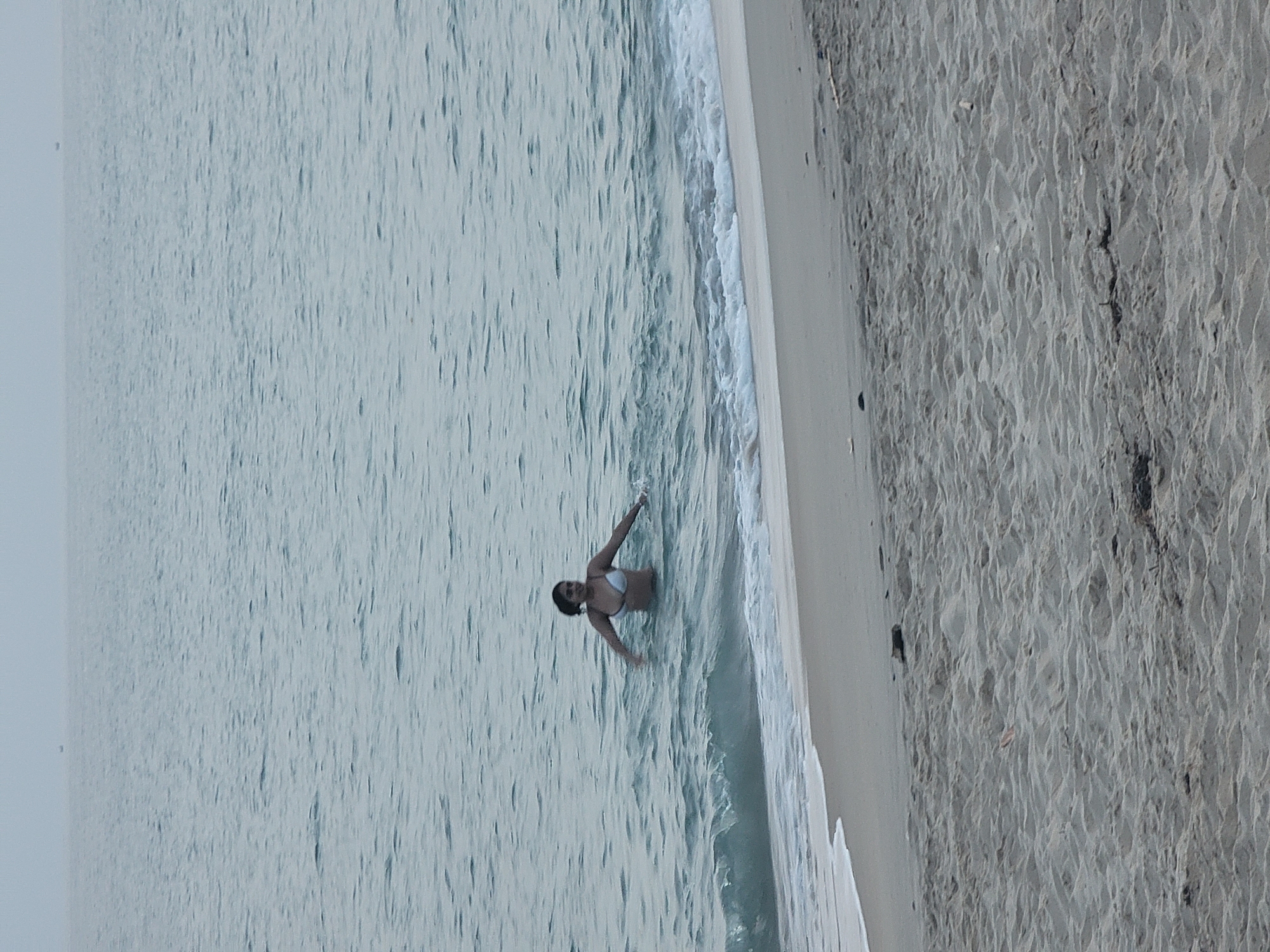 In the city of Quarteira, Portugal, a woman stands in the ocean, waist-deep in water. She is wearing a white bikini and has her arms outstretched. The water is calm with small waves breaking on the shore. The beach is sandy and appears to be mostly empty, with a few scattered pieces of debris. The sky is overcast, creating a muted light over the scene. In the distance, a small boat is visible on the horizon.